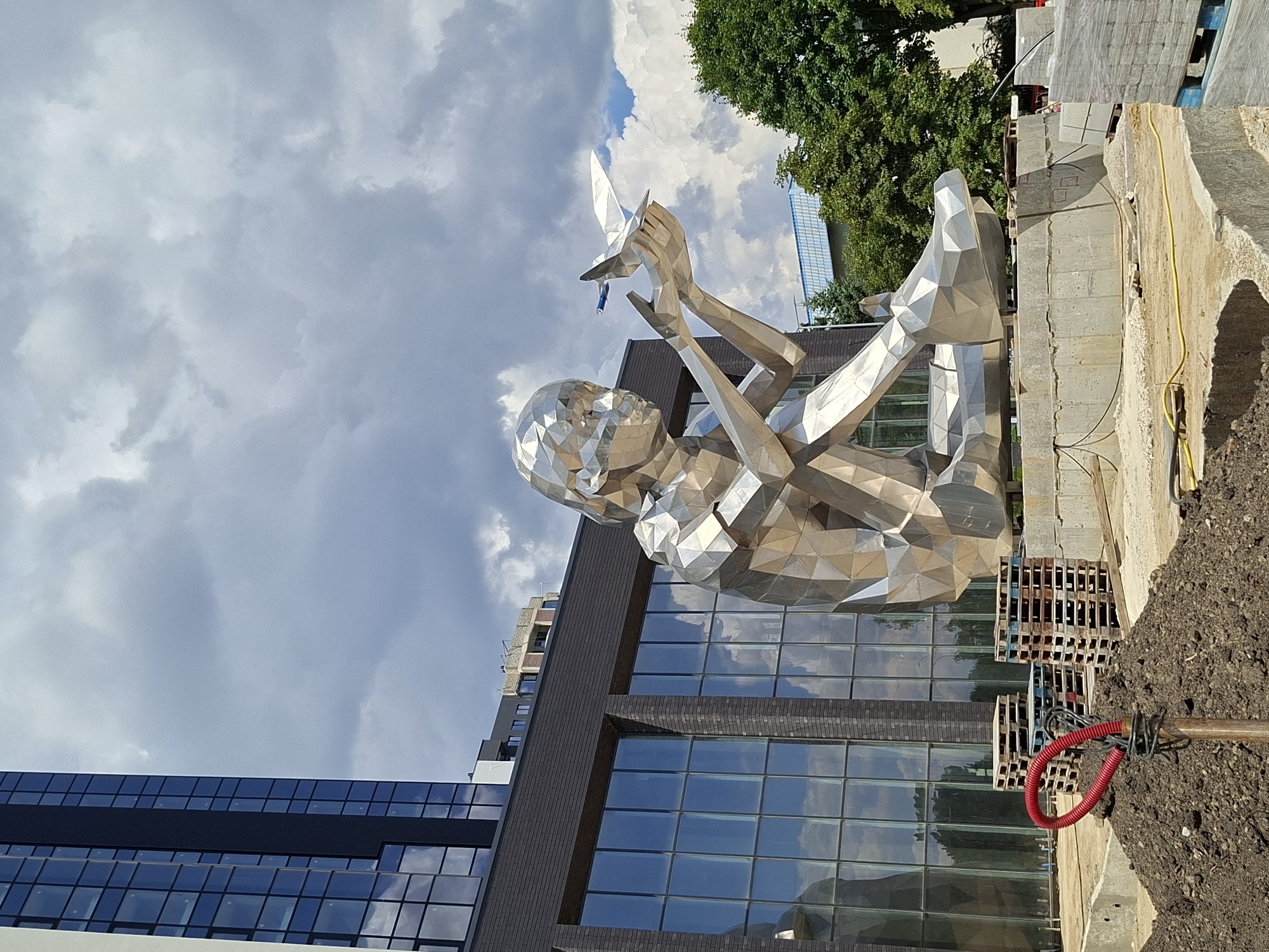 The image depicts a large, silver, polygonal sculpture of a seated child holding a paper airplane. The sculpture is positioned in an outdoor urban environment, possibly a construction site or public square. In the background, a modern, multi-story building with a dark facade and reflective windows is visible, as are trees and a cloudy sky. In the foreground, piles of soil and construction materials like wooden pallets and stacks of bricks are present, suggesting ongoing development or landscaping. A red hose is coiled on the soil. The sculpture is the central focus, with its geometric design and reflective surface catching the light. The overall scene suggests a contemporary urban setting with public art and development.