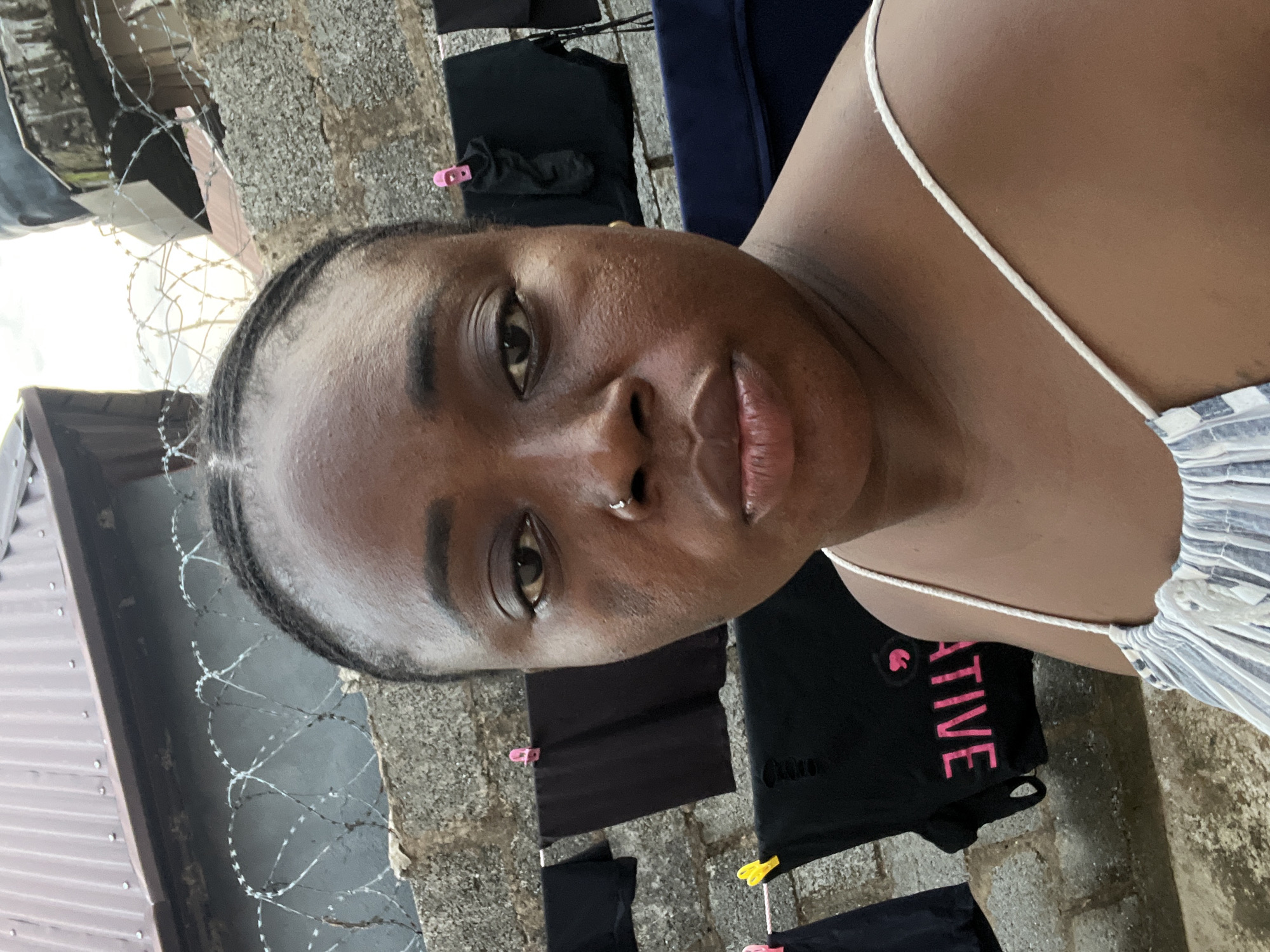 A close-up headshot of a woman with dark skin, likely in her early to mid-20s, is the central focus of the image. She has dark brown eyes, full lips, and her hair is styled in tight braids or cornrows. A small silver hoop is visible in her left nostril. She is wearing a light-colored top with thin white straps.
Behind her, clothes are hanging on a line to dry, secured with colorful clothespins. The backdrop appears to be a rough, textured concrete wall. To the left, barbed wire is visible against a corrugated metal structure, suggesting a boundary or security measure. The overall lighting is bright and natural, indicating an outdoor setting during the daytime.
Given the prevalence of clothing lines outside and the barbed wire, this image may be taken in a more residential or less affluent part of Abuja, Nigeria. However, without additional context, the exact location within Abuja cannot be determined definitively.