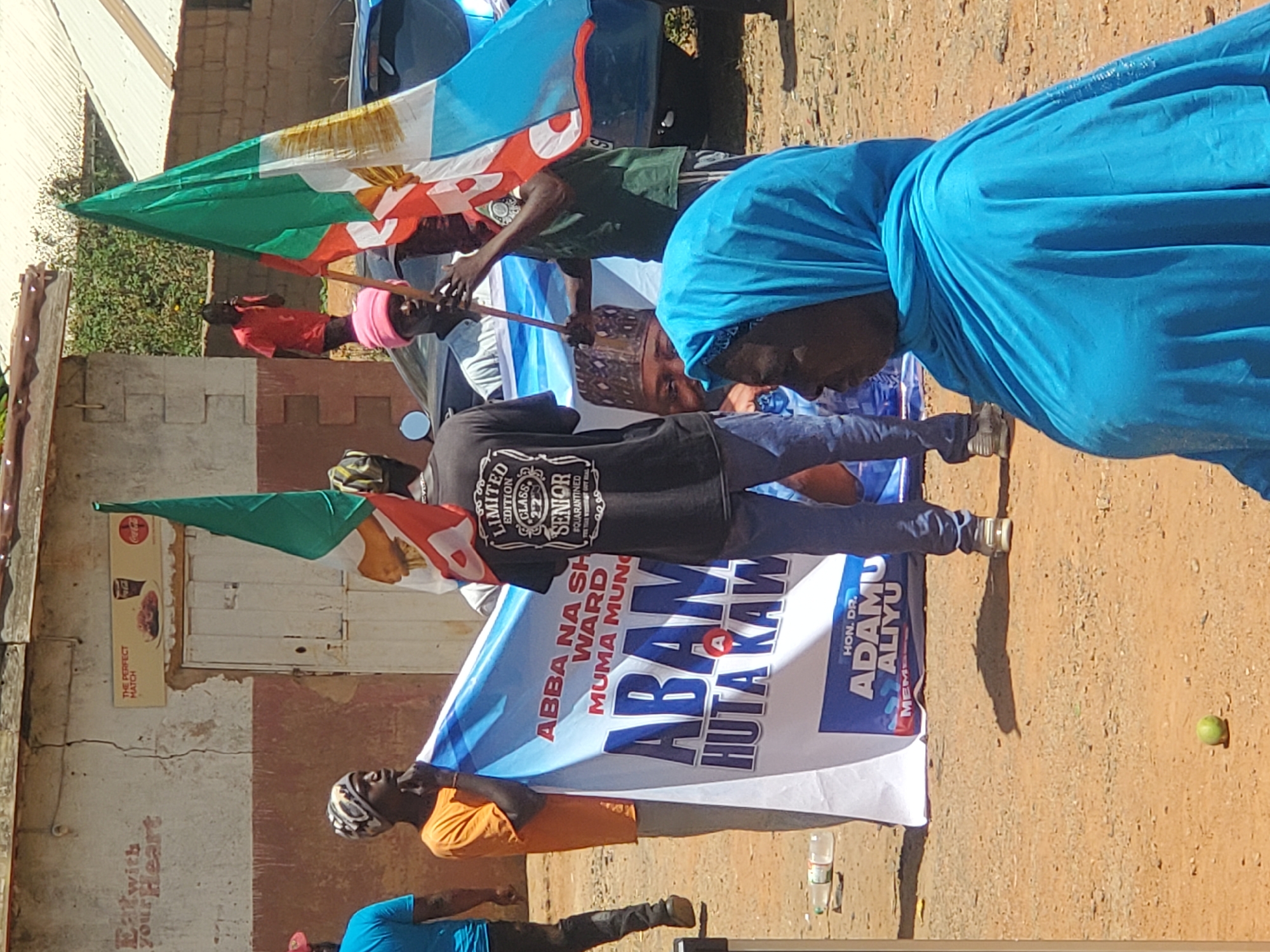 The image shows a group of people in what appears to be a political gathering or rally in Jos, Nigeria. People are holding Nigerian flags and a banner with political messages. The banner mentions "ABBA NA SH WARD MUMA MUNG" and "ABAM HUTA KAW," along with "HON. DR. ADAMU ALIYU." A woman in a blue headscarf is visible in the foreground. There is a Coca-Cola advertisement on a building in the background, with the words "Eat with your Heart" and "THE PERFECT MATCH." The ground is dusty, and a bottle of water and a green fruit are on the ground. One man wears a black shirt with "LIMITED EDITION CLASS SENIOR" printed on it.