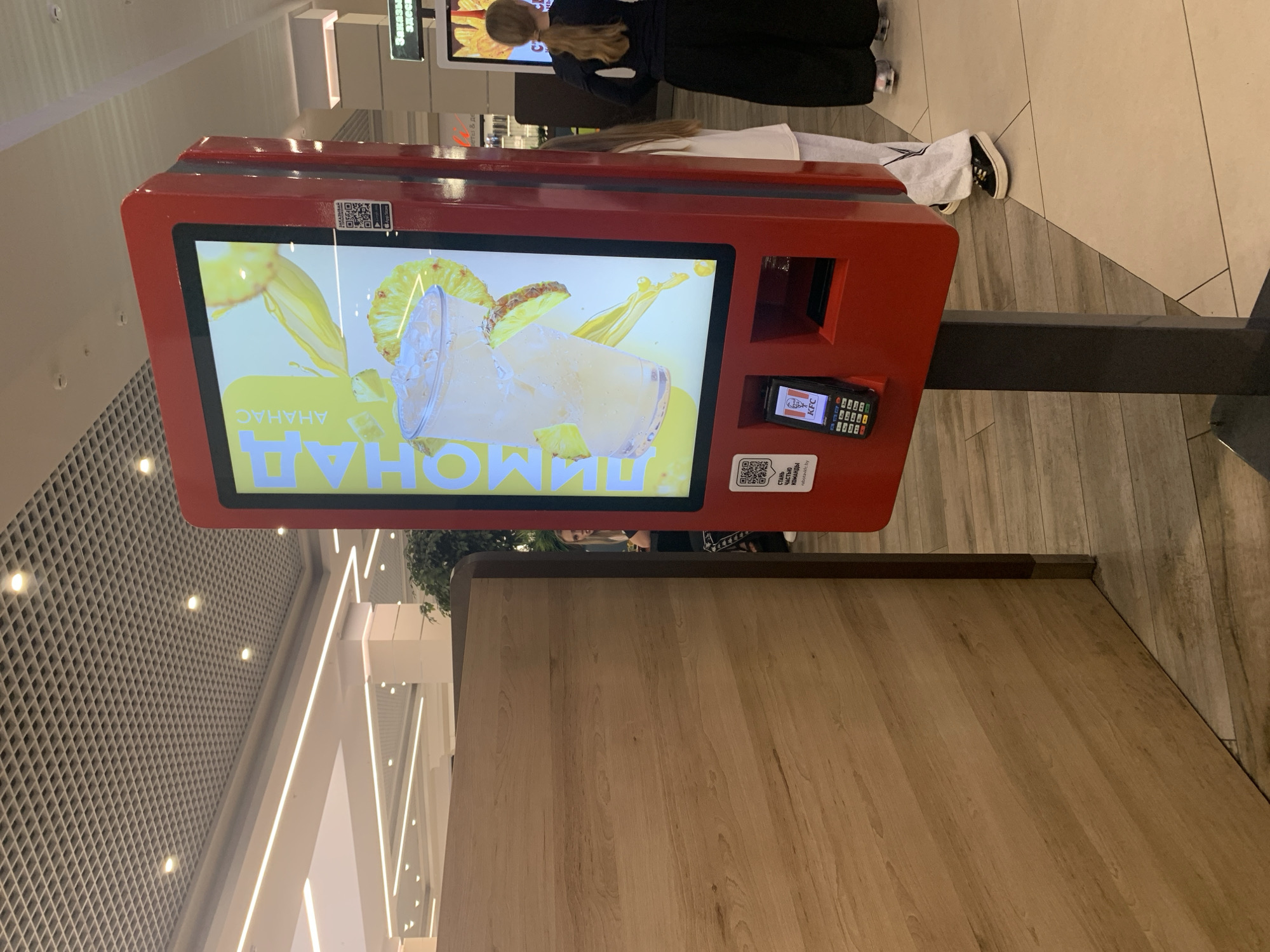The image shows a red KFC self-service kiosk in an indoor public space, likely a shopping mall, in Hrodna, Belarus. The kiosk's screen displays an advertisement for pineapple lemonade, with a large glass of the drink and pineapple slices. Below the screen, a payment terminal is visible, along with a QR code and app download instructions. To the left of the kiosk is a wooden partition. In the background, several individuals are present, including a woman with blonde hair in the middle ground, and two women in the background. One woman in the background is facing away from the camera and appears to be queuing or waiting. The floor is tiled with a light-colored pattern. The ceiling features a grid of acoustic tiles with recessed lighting.