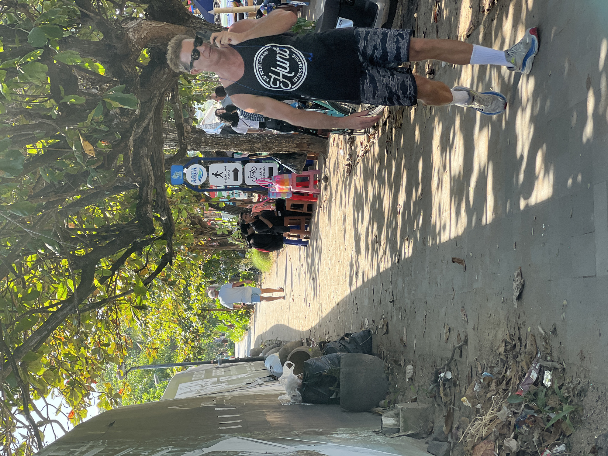 A man wearing a black tank top with the words "Hunt Keep Moving Forward" printed on it and camouflage patterned shorts walks along a sidewalk in Denpasar, Indonesia. He is speaking on a mobile phone. The sidewalk is lined with trees and foliage, and there is a concrete wall on the left side of the frame. Several trash bags and planters are piled against the wall. Further down the sidewalk, a man wearing a white t-shirt and blue shorts walks away from the camera. Several other people are visible in the background, as well as a sign with text in Indonesian and English. The ground is paved with gray stones, and fallen leaves are scattered on the path. The scene is brightly lit by the sun, creating dappled shadows from the trees.