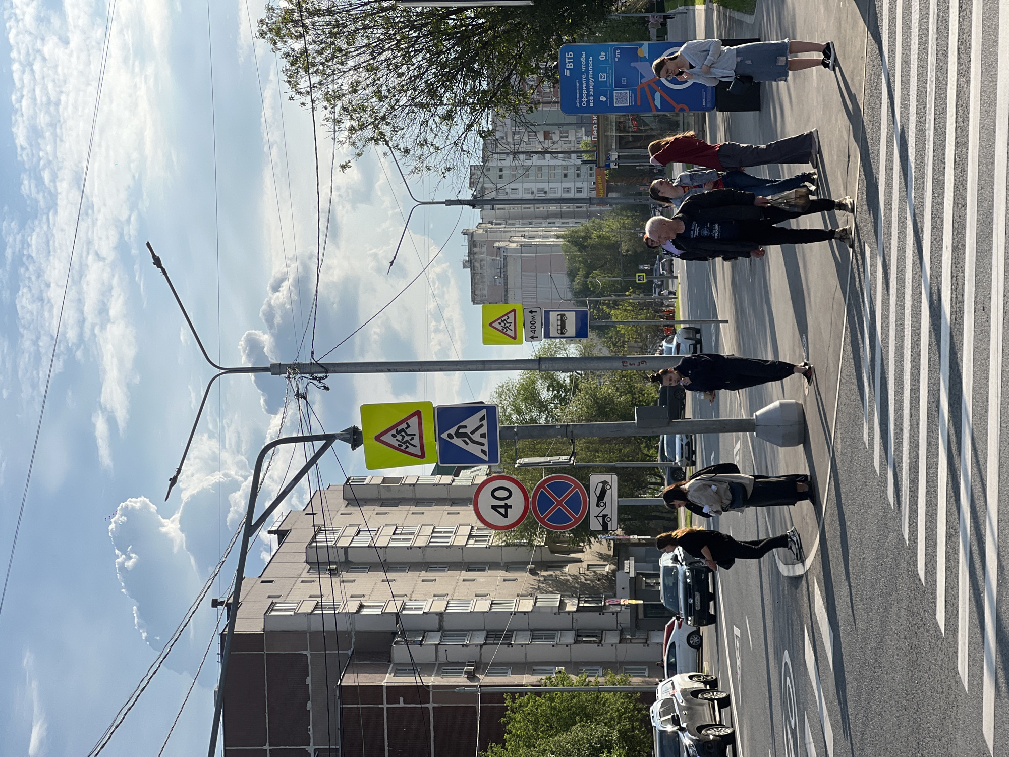 The image shows a street scene in Moscow, Russia. A group of pedestrians is crossing a crosswalk. The crosswalk is marked with white stripes. There are several traffic signs along the street, including speed limit (40 km/h), no parking, and pedestrian crossing signs. Buildings are visible in the background. A blue sign displaying information about VTB bank can be observed near the crossing.