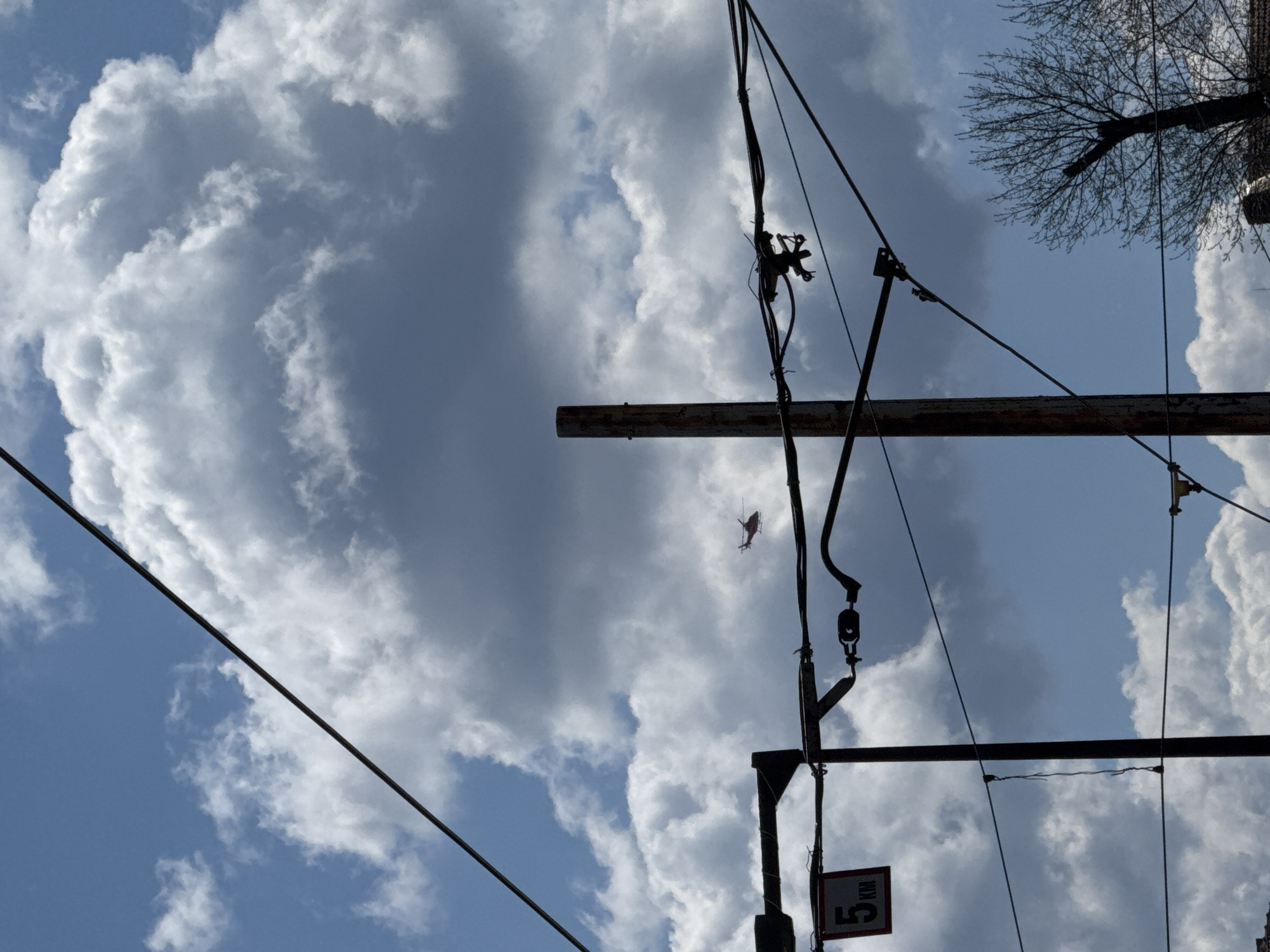 The image depicts a view looking upwards towards a partly cloudy sky, with utility poles and wires in the foreground. A helicopter is visible in the mid-ground between the utility structures and the clouds. Part of a tree is visible in the upper-right corner. A sign with the number "5" is attached to one of the utility poles. The location is Sofia, Bulgaria.