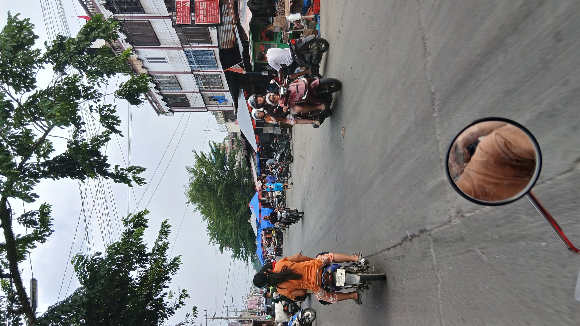 The image depicts a street scene in the city of Medan, Indonesia, likely a market area. The foreground is dominated by the rear view of a person riding a motorcycle, with a motorcycle mirror reflecting the rider's upper body. In the background, other motorcycles and riders are visible, along with several market stalls covered by blue and red awnings. People are seen walking and riding. Buildings with multiple windows line the right side of the street, some adorned with signs, including one for "META CELL." Lush green trees overhang the scene from above, and electrical wires crisscross the sky. The road surface is asphalt with visible cracks. The overall atmosphere suggests a bustling urban environment, typical of a marketplace in Medan.