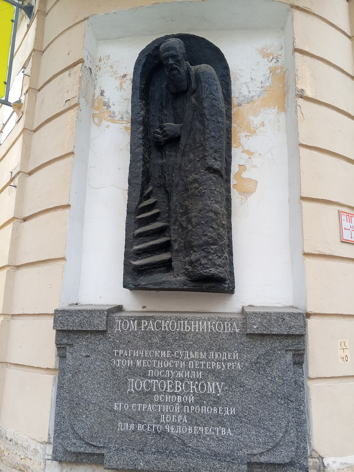 Here is a description of the image:
The image shows a dark, somewhat crude sculpture of a man standing in a niche above a gray granite plaque. The sculpture is of a figure with a slightly hunched posture and what appears to be a cloak or robe. A stairway seems to run next to the figure. The sculpture is mounted on an exterior wall of a building with a pale yellow façade.
Below the sculpture is a gray granite plaque with text in Cyrillic. The text is likely an inscription or memorial. A red and white sign, typical of those used to designate cultural heritage sites, can be seen attached to the wall to the right of the plaque.
The text on the plaque is partially translated as "Raskolnikov's House." This identifies the location as being connected to the character Raskolnikov from Fyodor Dostoevsky's novel "Crime and Punishment". The full inscription translates to "The tragic fates of people of this place in Saint Petersburg served Dostoevsky as the basis for his passionate sermon of good for all humanity."