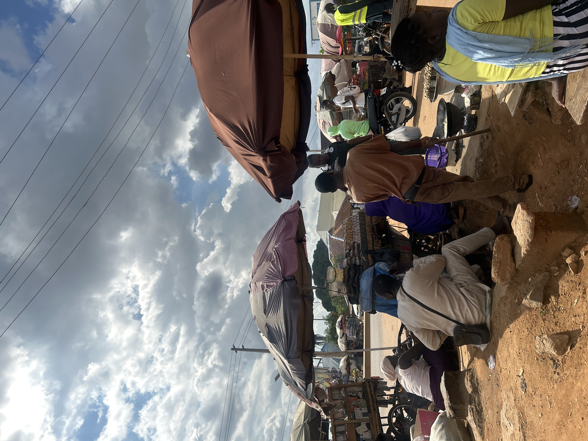 The image depicts a bustling street market scene in Rido, Nigeria, under a cloudy sky. Several people are present, some engaged in commerce, while others appear to be shoppers or bystanders. Large umbrellas, some faded and worn, provide shade for market stalls. One stall displays a variety of goods, including what appear to be packaged items and possibly food items. Another stall features baskets of produce, such as tomatoes and other vegetables, and what looks like garlic. A motorcycle is parked near one of the stalls, and a person in a high-visibility vest stands nearby. The ground is covered in reddish-brown dirt, with some stones and debris scattered around. A large black cooking pot is visible over a fire, suggesting food preparation is underway. The overall impression is one of vibrant local commerce and daily life unfolding in a Nigerian urban setting.