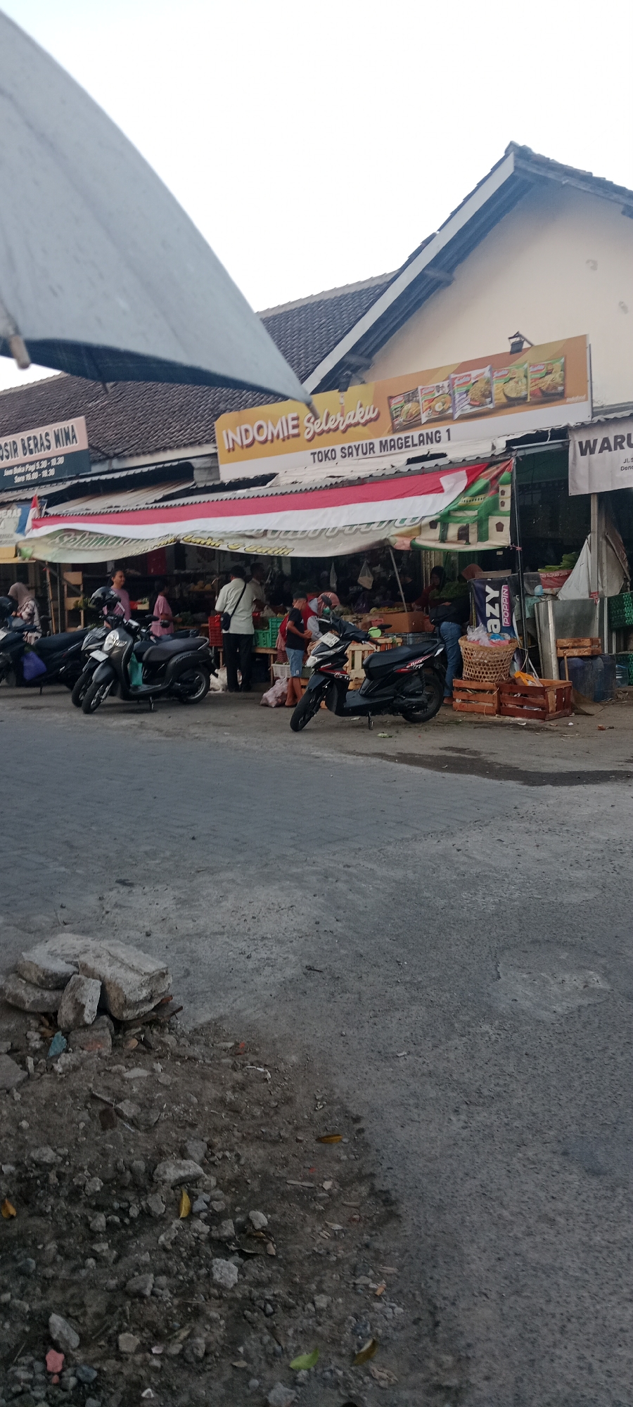 The image depicts an outdoor market scene in Maguwoharjo, Indonesia, likely in the afternoon given the lighting. In the foreground, a gray, cracked asphalt road is visible with a pile of construction debris and stones on the left side. A large, partially visible gray umbrella is angled from the upper left corner, casting a shadow.
Behind the umbrella, a row of market stalls and shops lines the street. A prominent orange banner with the text "INDOMIE SeleraKu" and images of Indomie noodle packets hangs above one of the stalls. Below this banner, another sign reads "TOKO SAYUR MAGELANG 1". A red and white Indonesian flag is draped across the front of this stall.
Several people are visible interacting within the market. Some are standing and observing goods, while others appear to be engaged in transactions. Various items are displayed for sale, including what seem to be vegetables and other produce. Scooters are parked in front of the stalls, suggesting this is a common mode of transportation. One scooter is positioned prominently in the middle of the frame. To the far right, a sign for "WARU" is partially visible, and crates and baskets are stacked. The overall environment appears to be a bustling, informal marketplace.