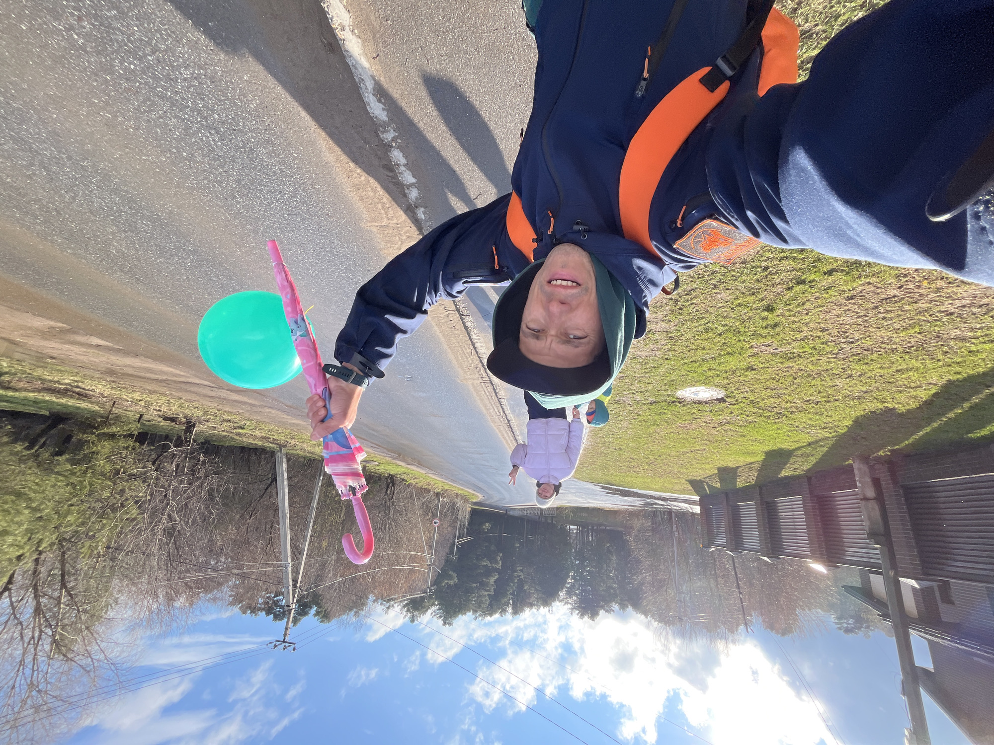 The image depicts a man taking a selfie on a road. He is holding a green balloon and a pink umbrella. In the background, a child in a white coat is walking. The surrounding environment includes trees, grass, buildings, and power lines. The location is Chaikovsky, Russia.