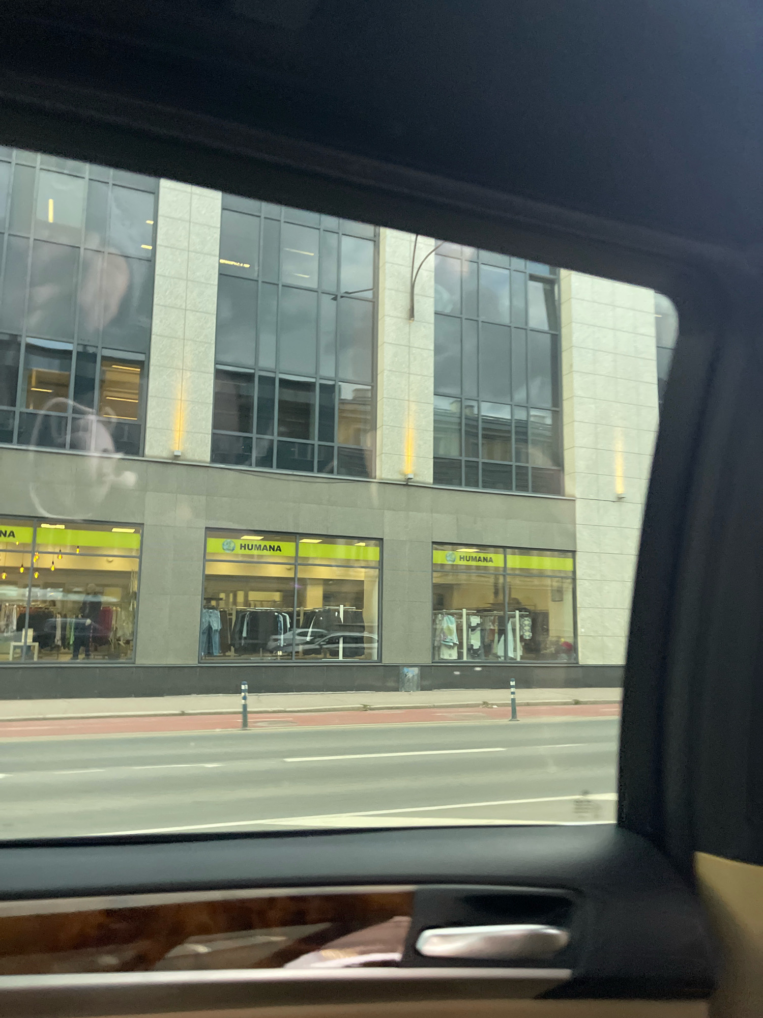 This is a photograph taken from inside a vehicle, looking out at a street scene in Tallinn, Estonia. The focus is on the facade of a multi-story building with large windows. The building's exterior is a light-colored stone or concrete, with darker framed windows. Below the upper windows, there are two ground-level storefronts with bright yellow signage. The left storefront has "HUMANA" visible on its sign, and the right storefront also displays "HUMANA" with a green circular logo. Both storefronts are illuminated and display clothing racks and mannequins, indicating they are retail clothing stores. The street in front of the building is paved with asphalt and has red and white lane markings. There are a few traffic signs and bollards visible on the sidewalk. The interior of the vehicle is partially visible in the foreground, including a car door handle and trim. The overall scene suggests an urban environment with commercial activity.