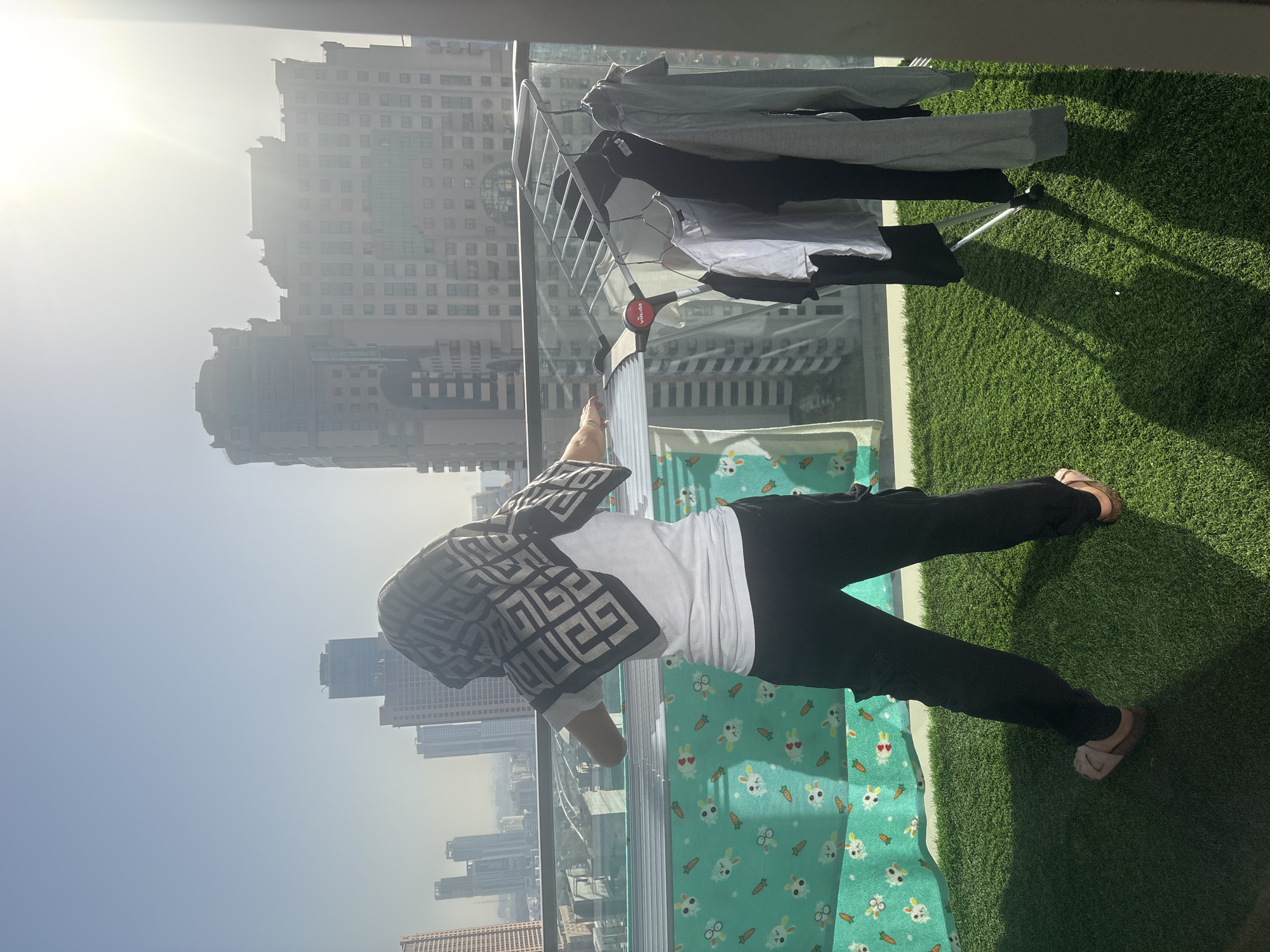 A person stands on a balcony with artificial turf flooring, facing away from the camera and towards a cityscape. The person is wearing a white t-shirt, black pants, and a patterned headscarf. They are positioned next to a drying rack with several clothes hanging on it, including shirts and a sweatshirt. Behind the person, tall buildings, characteristic of Dubai, United Arab Emirates, rise against a bright sky with visible sunlight and haze. The balcony has a glass railing. A green fabric with a white bunny and carrot pattern is visible to the left of the person. The sun casts long shadows on the artificial turf.
