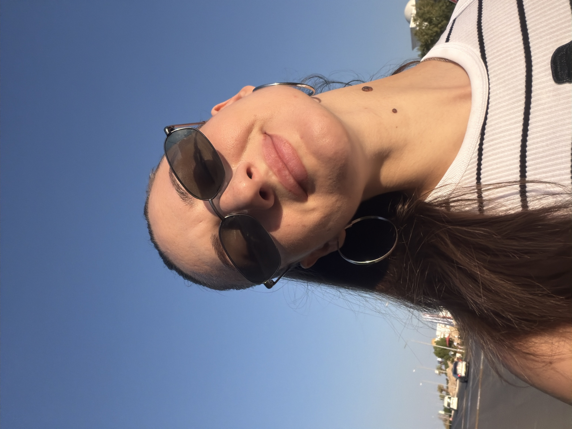 A young woman is pictured in a close-up, upward-angled selfie taken outdoors. She is wearing sunglasses and a white and black striped t-shirt. She has long brown hair and large hoop earrings. There are two moles on her neck. The background shows a clear blue sky and a street with cars, streetlights, and some buildings, suggesting an urban or suburban environment. This scene is consistent with locations in Sharm El Sheikh, Egypt, where such a clear sky and road infrastructure are common.