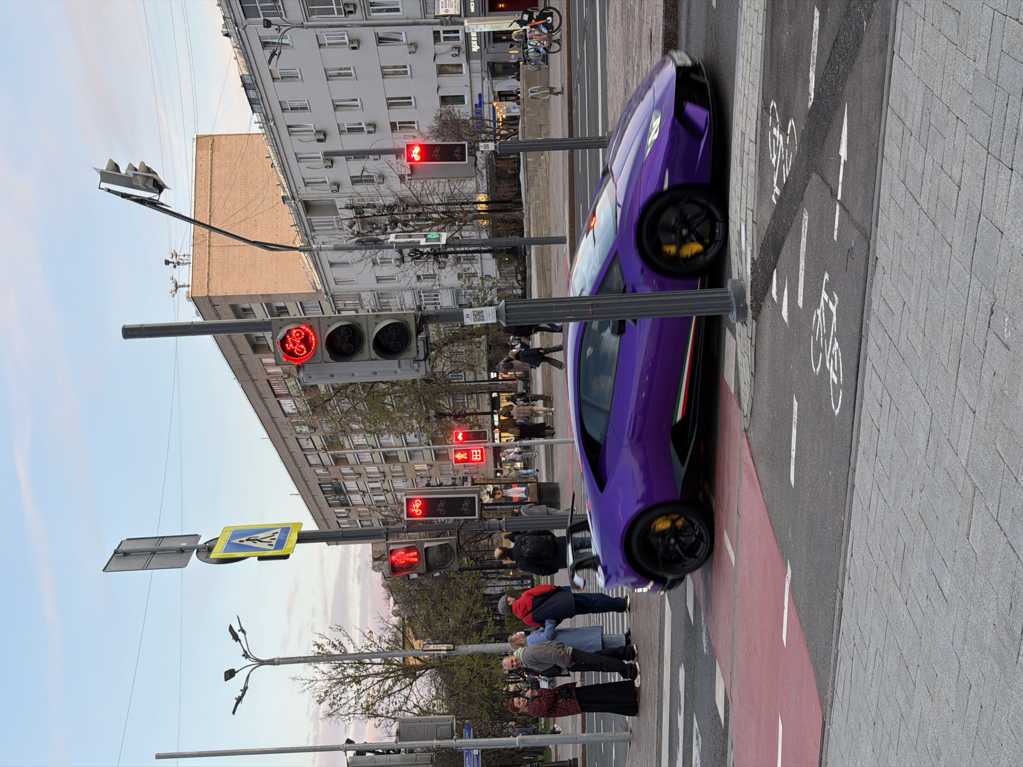 The image depicts a street scene in Moscow, Russia. A purple Lamborghini sports car is parked on a red-painted bicycle lane next to a crosswalk, positioned on the right-hand side of the frame. Several pedestrians are standing near the crosswalk, including a group of elderly individuals. Traffic lights are visible above the crosswalk, displaying a red light for bicycles. The surrounding buildings appear to be apartment complexes and commercial establishments. The street itself is paved with a combination of asphalt and grey brick. A pedestrian crossing sign is placed on a pole. The sky is partially visible in the background, with a light blue color. A bicycle lane sign is visible on the ground.