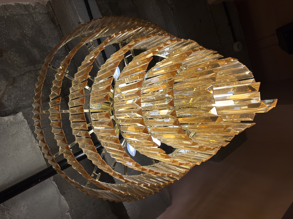 The image displays a multi-tiered chandelier, constructed from gold-toned metal and numerous amber-tinted crystal or glass prisms. The prisms are arranged in a spiraling, inverted conical formation, with each tier progressively smaller towards the bottom, creating a pointed base. Visible incandescent light bulbs are present within the chandelier's structure, illuminating the prisms. The chandelier is suspended from a raw, exposed concrete ceiling, showing cracks and irregular textures. Overhead, dark pipes or conduits are visible against the concrete. The background walls are a dark, reddish-brown hue. In the lower right, a dark, rectangular speaker box is mounted, and a circular white fixture, possibly a smoke detector or sprinkler head, is visible on the wall further back. There are no individuals present, nor are any actions or interactions depicted. The visual information within the image does not provide identifiable markers for a specific location within Tel-Aviv, Israel.