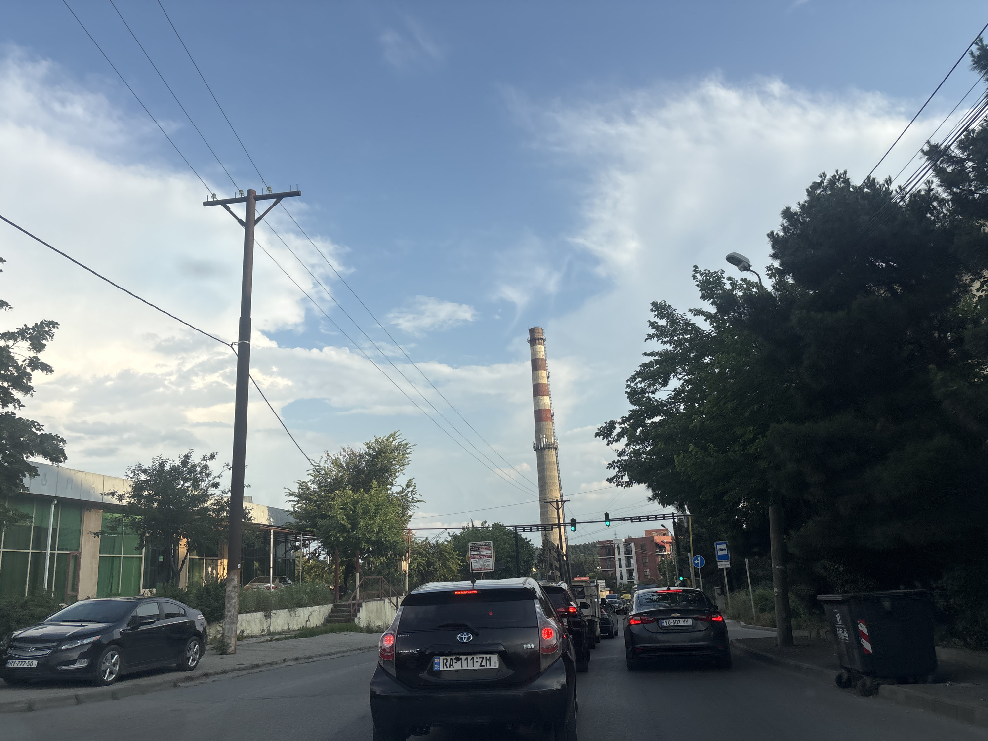 The image depicts a street scene in Tbilisi, Georgia, likely during daylight hours with overcast skies. The foreground shows a traffic jam with several cars on a paved road. The lead vehicle is a black hatchback with a Georgian license plate that reads "RA-111-ZM". To its left is a black sedan with a license plate "RY-777-SD". Further back, other vehicles are visible, including a dark gray sedan with a license plate "YU-607-VV".
Along the right side of the road, there are large, dark green trees and a black trash receptacle. A concrete sidewalk is visible between the road and the trees. On the left side of the road, there is a building with a glass facade and green panels. Adjacent to this building is a bare-bones concrete structure with metal supports, possibly a bus stop shelter.
In the middle ground, a tall, striped industrial chimney with red and white bands stands prominently against the sky. Power lines and utility poles are a significant feature throughout the scene. A street light is visible among the trees on the right. In the background, more trees and buildings, including apartment blocks, can be seen. The overall impression is a typical urban environment experiencing traffic congestion.