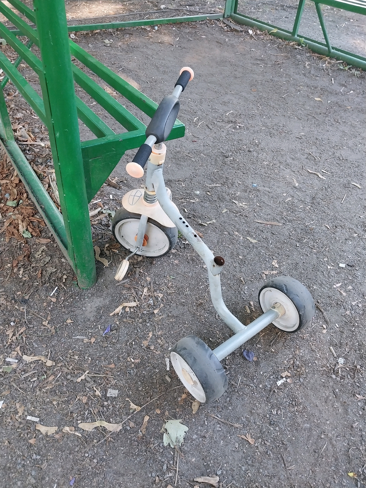 A disused child's tricycle with a faded orange and white frame lies on its side on a dirt and gravel surface. The tricycle is near a green metal bench and a green metal fence. The ground is littered with dry leaves and small debris. The scene appears to be outdoors in an urban or park-like setting. The tricycle has three wheels, handlebars with orange grips, and a pedal. The background shows more of the green fence and dappled sunlight filtering through trees. The overall impression is one of neglect and abandonment.