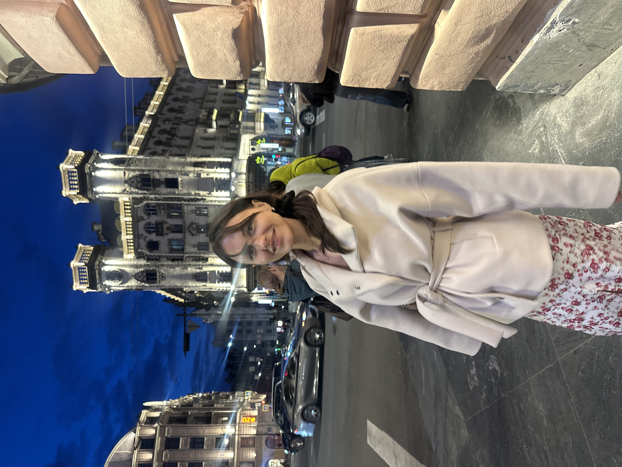The image depicts a woman in Saint Petersburg, Russia, at night. She wears a light-colored coat over a floral dress. The background features illuminated buildings with intricate architecture, typical of the city's historic center. Cars are parked and driving on the street. The sky is a dark blue, indicating the late hour. A bridge or an elevated structure is visible overhead. The woman is standing on a paved surface, possibly a sidewalk or pedestrian area.