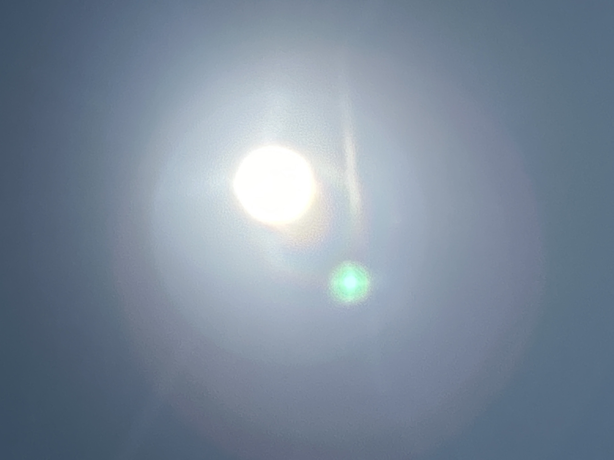 The image shows a bright, overexposed sun in a hazy sky, creating lens flare and a green light artifact to the left of the sun. The sky is a gradient of light blue to white. There is no discernible city, environment, people, or action depicted. The location in Matraevo, Russia, is not visible.