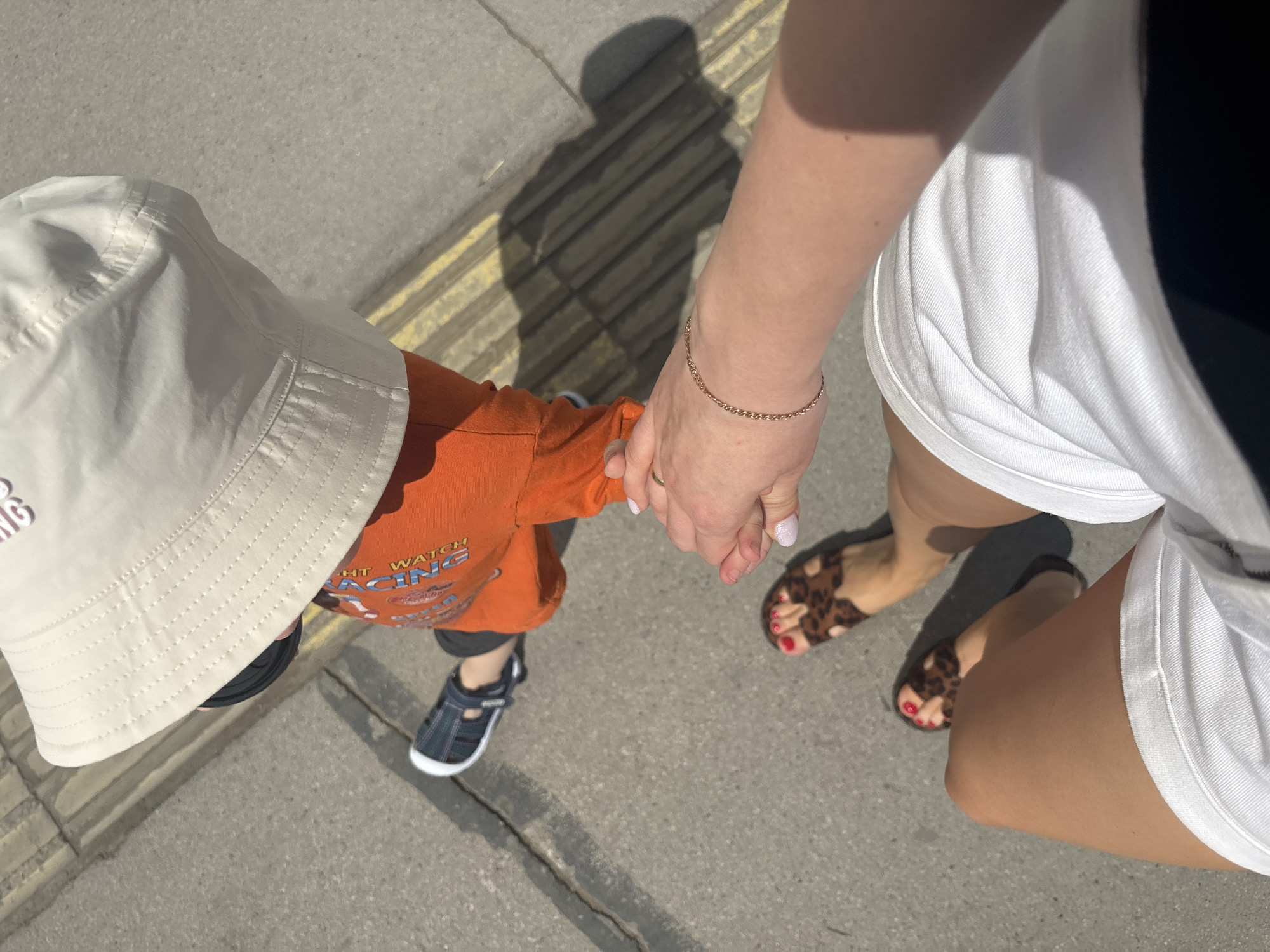 The image shows a person holding the hand of a toddler on a sidewalk. The toddler wears a beige bucket hat, an orange t-shirt with a racing-themed design, dark-colored shorts, and navy blue sandals. The person holding the toddler's hand is wearing a gold bracelet and a white dress or skirt over black shorts. They are wearing leopard-print sandals, and their toenails are painted red. The sidewalk is concrete with yellow tactile paving markings. The image is taken from an overhead perspective, casting a shadow of the subjects on the ground. While there are tactile paving markings, the image does not provide sufficient information to confidently confirm the location is Perm, Russia.