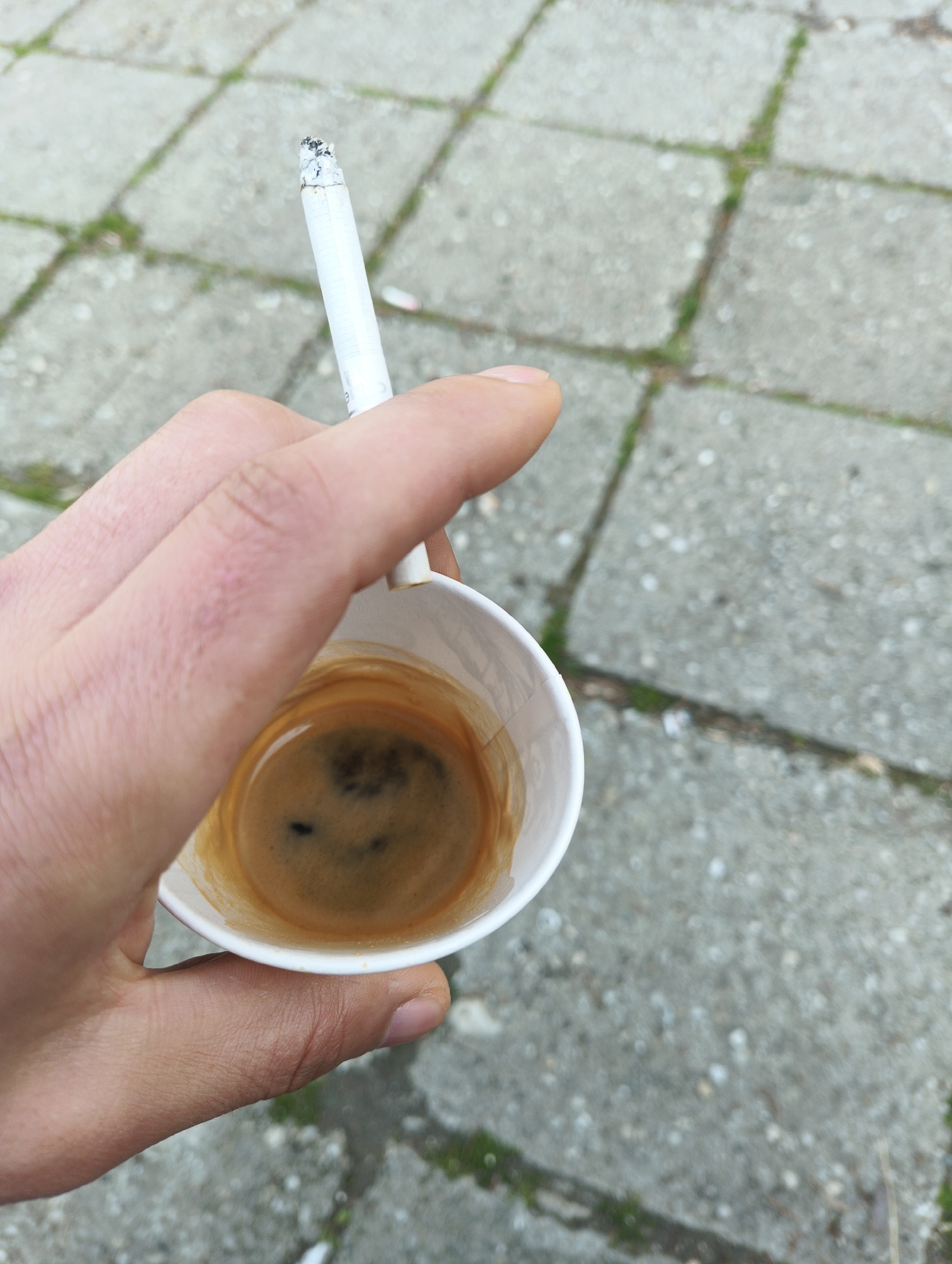 A first-person, high-angle shot depicts a hand holding a white, disposable cup filled with dark coffee and a lit cigarette. The hand appears dry, possibly weathered. The background consists of a sidewalk composed of light gray concrete tiles, interspersed with thin lines of green moss or grass. The location is not identifiable.