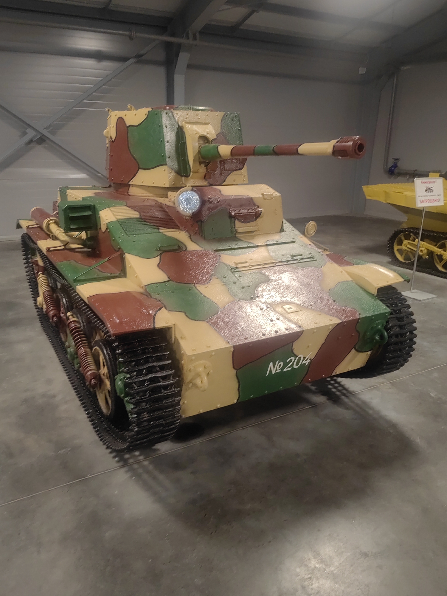 An overhead shot shows a World War II-era Soviet BT-7 tank on display in a museum. The tank is painted in a camouflage pattern of tan, green, and brown. The hull is numbered "№ 204". In the background, to the right, is a yellow tracked vehicle and a sign. The museum appears to be a large, industrial-looking building with a corrugated metal roof and grey walls. The floor is concrete. The lighting is typical of an indoor museum exhibit.