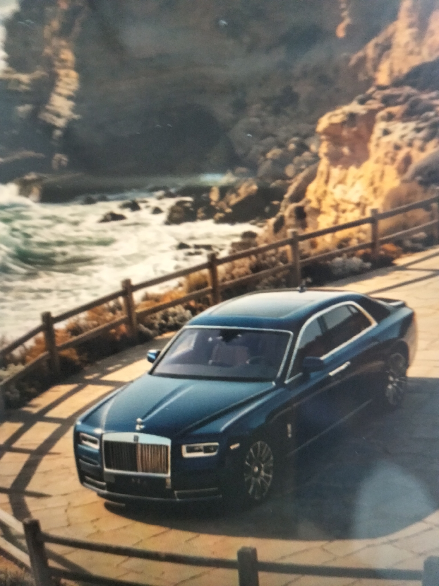 A dark blue Rolls-Royce sedan is parked on a paved surface next to a wooden fence. The car is facing towards the left of the frame. In the background, there is a body of water with waves crashing against a rocky shore, and cliffs with vegetation. The sky is overcast. The scene is bathed in natural light. The location is identified as Payagaboh, Indonesia, a coastal area characterized by its natural beauty, including cliffs, beaches, and ocean views. The presence of the luxury car suggests a possibly scenic viewpoint or a high-end resort.