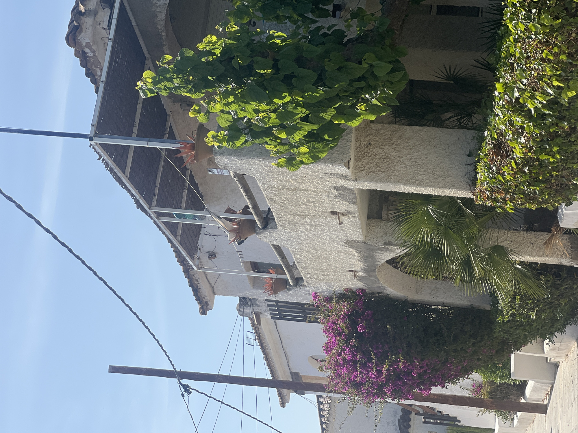 The image depicts a two-story building located in La Marina De Creixell, Spain. The building has a white, textured exterior with a tiled roof. Lush green vegetation, including bougainvillea with vibrant purple flowers, is growing around the building. Electrical wires are visible in the foreground, running diagonally across the blue sky. The architecture and vegetation suggest a Mediterranean environment.
