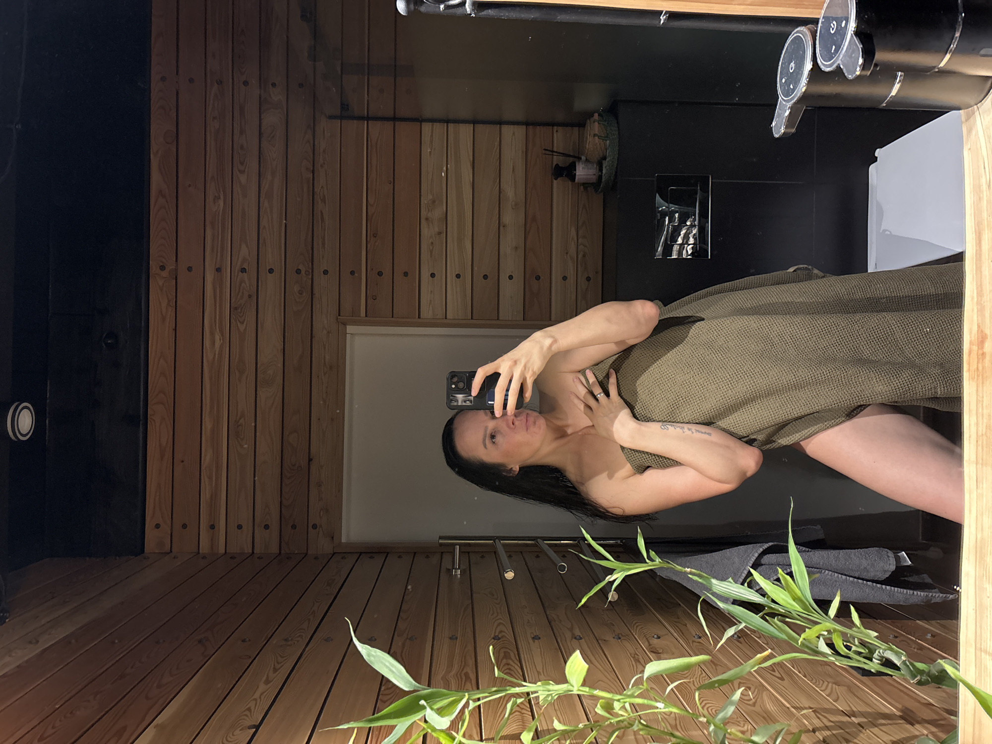 A woman stands in a wood-paneled sauna, holding a cell phone. She is partially wrapped in a green towel and is taking a selfie, facing a mirror. Bamboo plants are placed in the foreground, and a towel rack is located behind her. The wall paneling indicates the location may be within a private sauna facility. The city of Chaikovsky, Russia, is not directly discernable from the image.