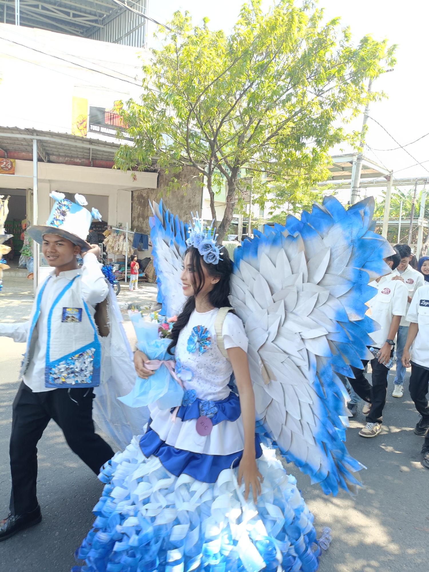 The image captures a vibrant outdoor scene, likely a parade or festival, in Kediri City, Indonesia. In the foreground, a young woman is dressed in an elaborate costume featuring large, white and blue feathered wings and a multi-layered blue and white skirt. She holds a bouquet wrapped in light blue paper. Beside her, a young man, also in costume, wears a tall, wide-brimmed hat adorned with blue accents, a white shirt, and a decorative vest with blue detailing. He is adjusting his hat. Behind them, other individuals, some in white shirts, are visible, suggesting a procession. The background shows buildings with awnings, a tree with lush green leaves, and electrical wires, indicative of an urban street setting. The overall atmosphere appears festive, with bright sunlight casting shadows on the ground.