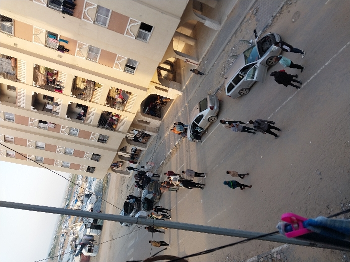 The image presents an elevated view of a street scene in Khan Yunis, Palestinian Territory. On the right, a multi-story, light-colored residential building features numerous windows and balconies with decorative railings, some displaying hanging laundry. The ground floor has arched open passages. A paved, somewhat dusty street occupies the middle ground. Scattered across this street are approximately two dozen individuals, predominantly males and youths, dressed in casual clothing, walking or standing in small clusters. Two silver/grey vehicles are visible on the right side of the street: a hatchback with its driver's side door open and a station wagon with its hood raised and multiple passenger doors ajar. A third dark-colored vehicle with an open rear compartment is further up the street on the left. To the left of the street and beyond an unpaved area, an extensive makeshift camp consisting of numerous white and grey tents and temporary shelters stretches into the background, with distant hills visible under a clear sky. A utility pole with wires runs vertically in the foreground on the left, from which a pink and blue garment hangs.
