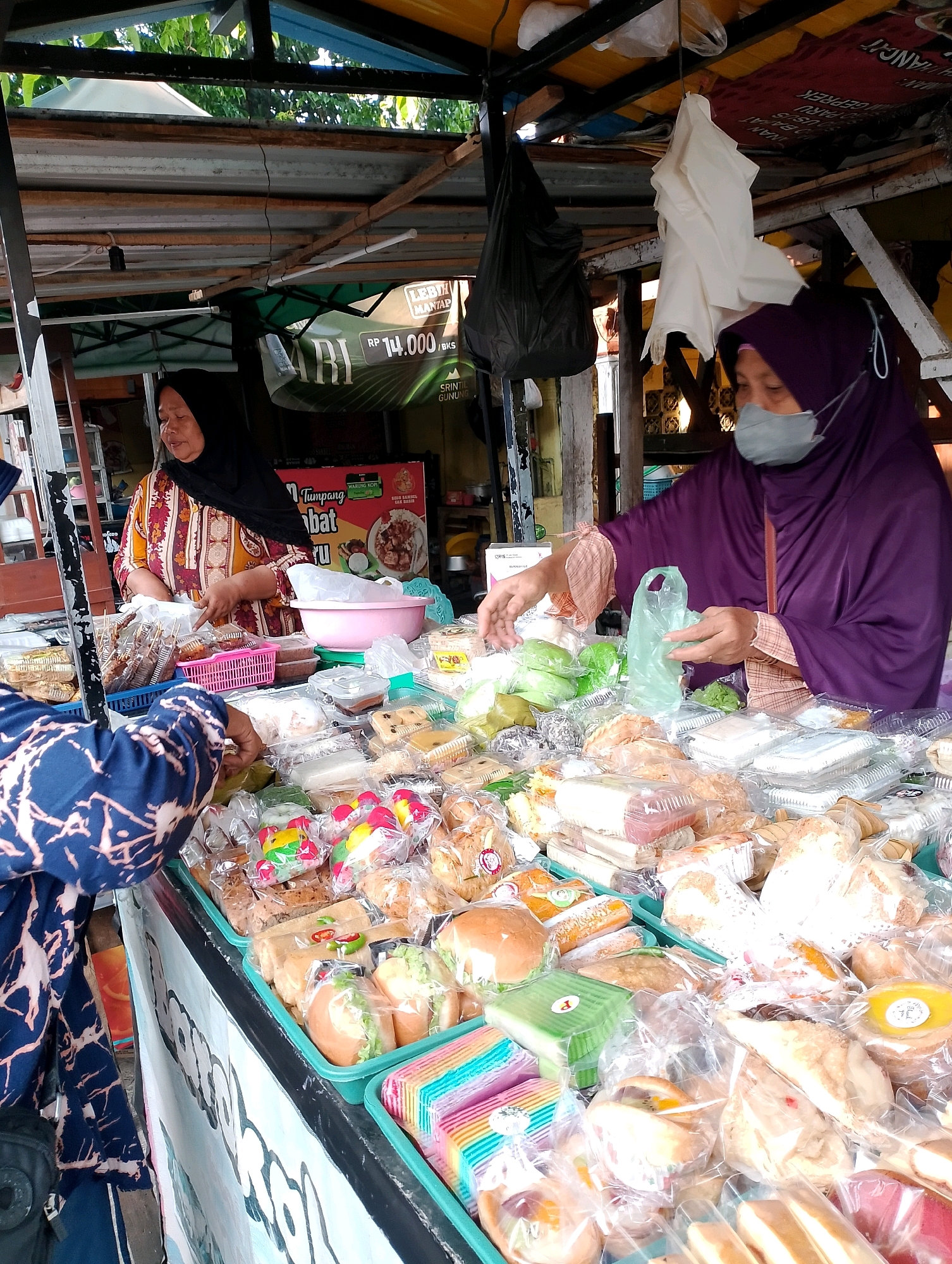 The image captures a street food stall in Probolinggo, Indonesia, overflowing with various packaged food items, likely snacks and baked goods. Two women are visible, appearing to be vendors. The woman on the left, wearing a black hijab and a colorful patterned top, stands behind a counter laden with food. The woman on the right, wearing a purple hijab and a face mask, is actively engaged with the stall, holding a plastic bag and reaching for more items. The stall itself is under a makeshift shelter with a corrugated metal roof. The display is densely packed with goods in plastic packaging, including bread, pastries, and what appear to be rice cakes or sweet treats. The background hints at an outdoor market setting with trees and other structures visible.
