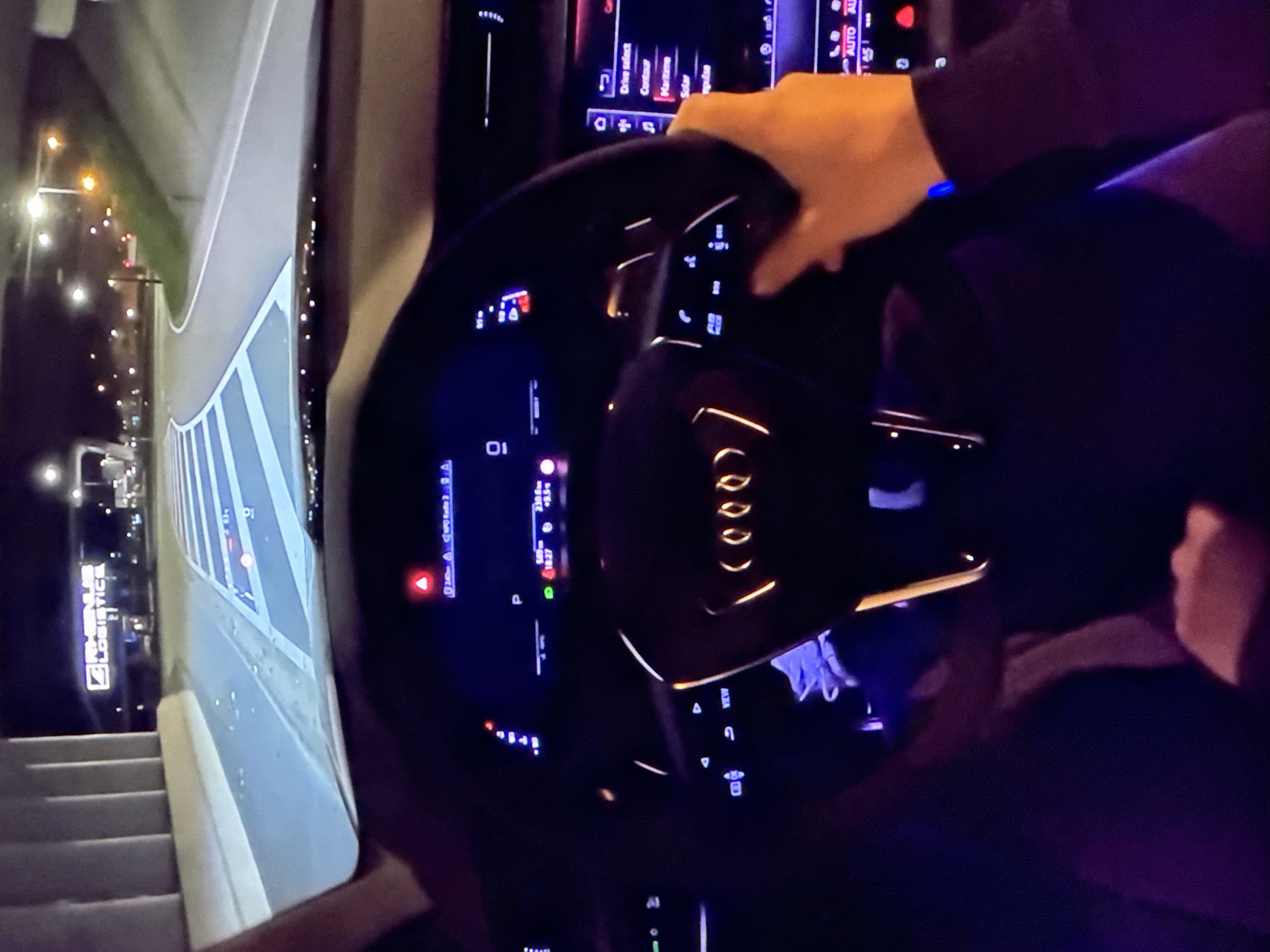 The image depicts the interior of a car at night, with the driver's perspective focused on the steering wheel and dashboard. The steering wheel, with the Audi logo illuminated in orange, is partially obscured by a hand. The dashboard displays a variety of illuminated readouts, including speed, gear selection, and a navigation interface. Outside the car, the road is visible, marked with blue and white lines, indicating a highway or major road. In the distance, illuminated signs for "RHENUS LOGISTICS" are visible, suggesting an industrial or logistics hub. Streetlights and building lights dot the background, illuminating the surrounding environment. The scene appears to be in Bezden, Bulgaria, based on the context provided.