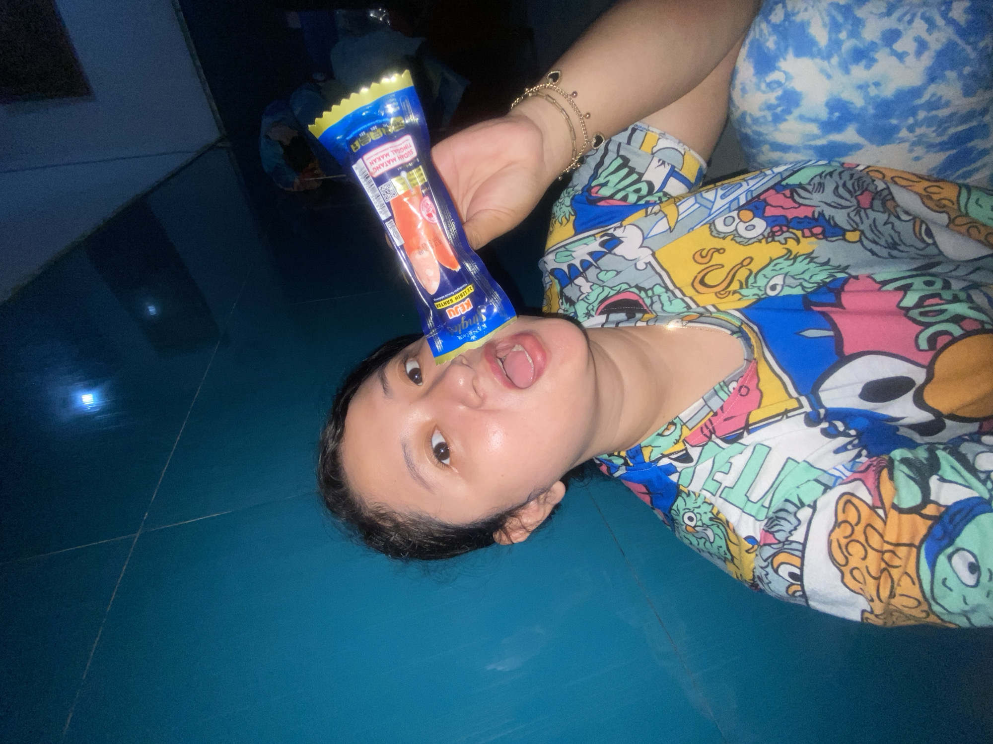 An indoor scene in Gelam, Indonesia, depicts a young adult female looking directly at the camera. She wears a multi-colored t-shirt patterned with cartoon characters and blue and white tie-dye patterned pants. A gold-colored, multi-strand bracelet with small dark heart-shaped charms is on her right wrist. She holds a blue and yellow flexible plastic food package, containing two pinkish-orange sticks and marked with Indonesian text including "SIAP MATANG TINGGAL MAKAN" (Ready to eat), horizontally towards her open mouth. The background is a plain teal-blue wall, with a faint light source or reflection visible in the upper right quadrant.