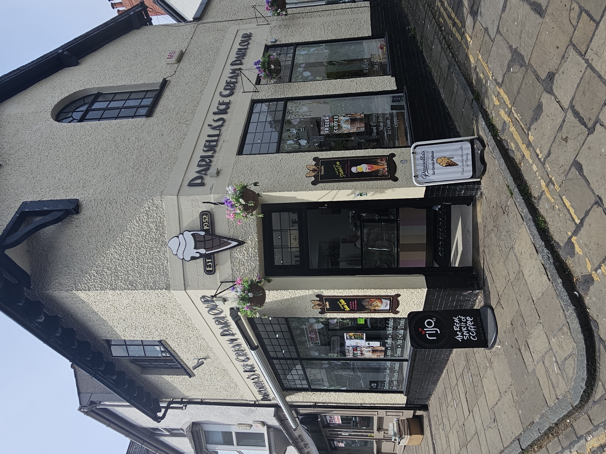 The image shows the exterior of Parisella’s Ice Cream Parlour, located in the city of Conwy, United Kingdom. The building has a cream-colored textured facade with black trim around windows and doors. The parlour has two large display windows, with advertisements for frozen coffee and ice cream products. A sign with an ice cream cone logo marks the establishment's founding year, 1952. Sidewalk sandwich boards advertise Parisella's ice cream and rijo coffee. A sign on one of the sandwich boards advertises ice cream made within the walls of Conwy. The street is paved with light-colored stone and has yellow double lines marking the curb. The building appears to be situated on a corner.