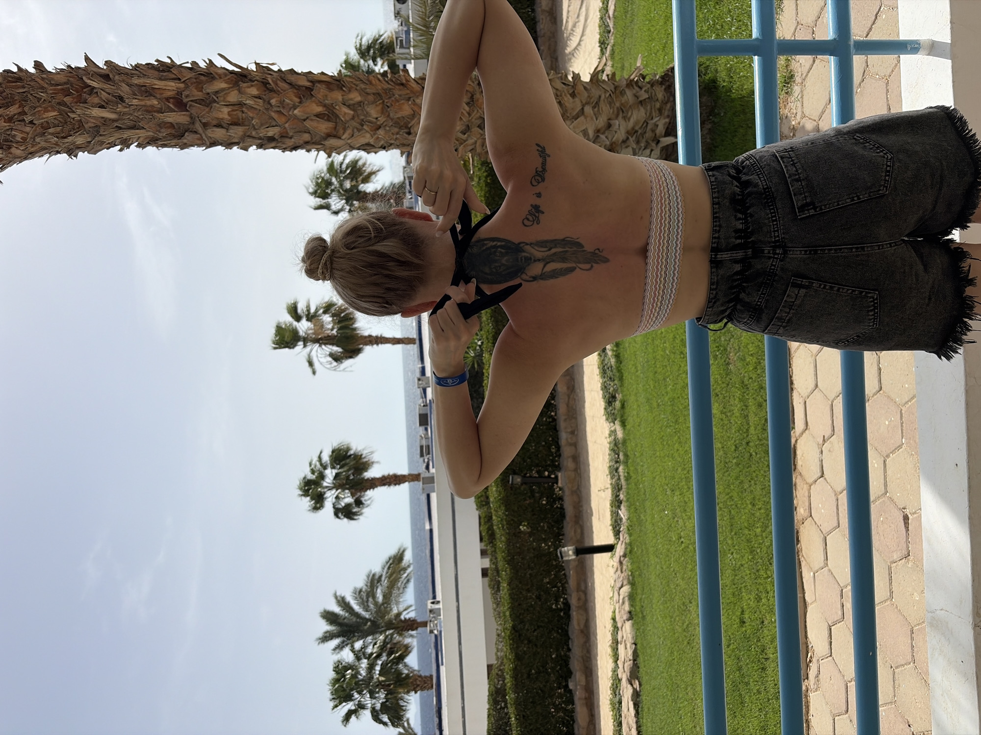A medium, horizontally-oriented shot depicts a woman from behind with her back tattoos in view. She is on a balcony with blue railings, with grass and palm trees visible in the background. The sky is light blue with some clouds, and the location appears to be Sharm El Sheikh, Egypt.
The woman has her hair up in a bun and is wearing dark gray denim shorts with a fringed hem. She has a tattoo on her back consisting of two parts: a dreamcatcher with feathers, and the phrase "Life is Beautiful" written above it. She is adjusting a black garment around her neck. She is also wearing a blue bracelet.
The environment includes palm trees and a building in the background, as well as the balcony, which has a paved floor. There is also some greenery visible between the balcony and the trees.