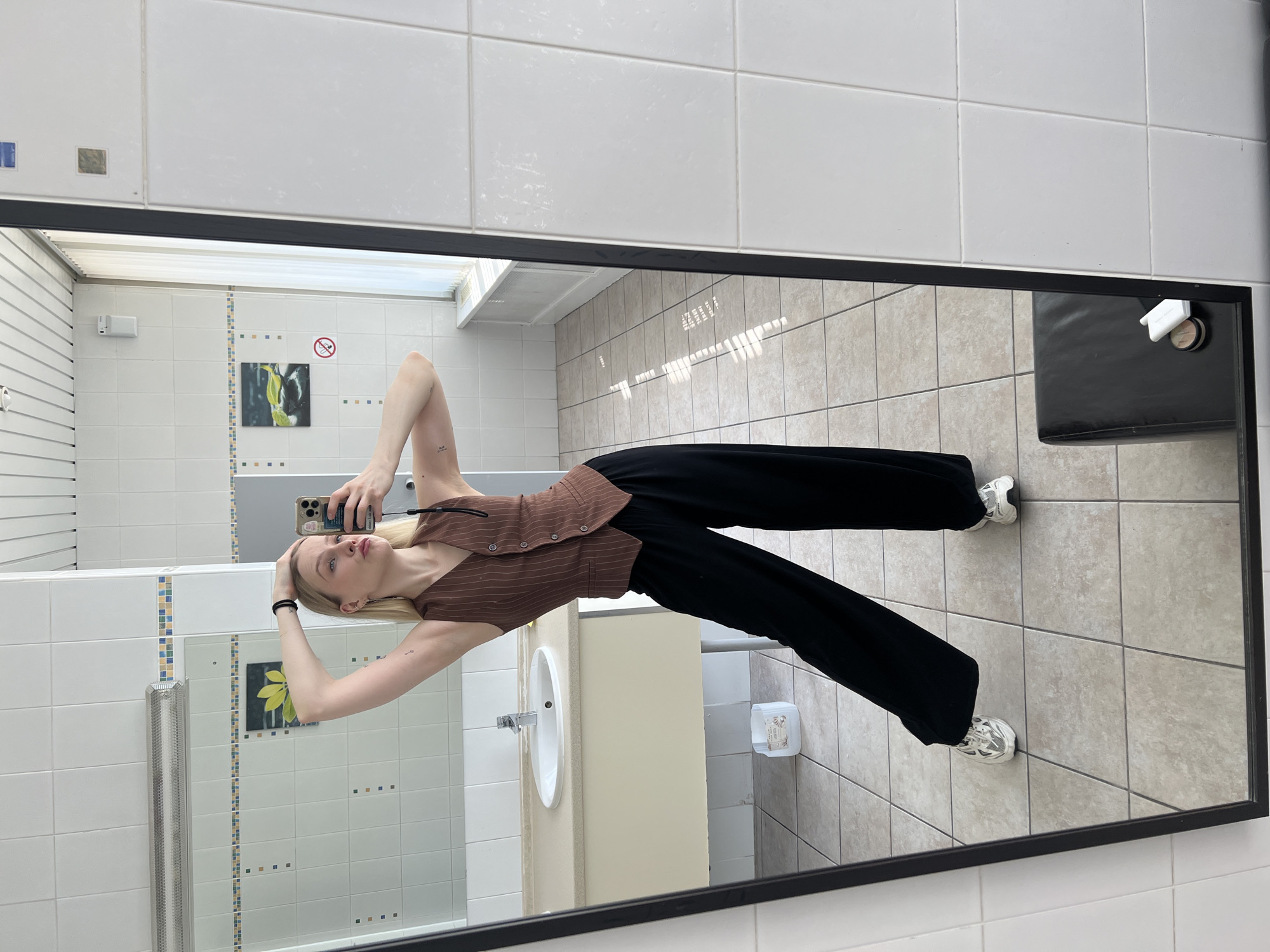 A woman is taking a picture of herself in a mirror. The mirror is in a bathroom with white tiles and beige floor tiles. The woman is wearing a brown pinstripe top and black pants. Her hair is blonde and pulled back with a black hair tie. She is holding a smartphone in her right hand. On the right side of the mirror, a black seat is visible. The bathroom includes a sink with a white base and a chrome faucet, plus an electrical switch. The location is the city of Khimki, Russia.