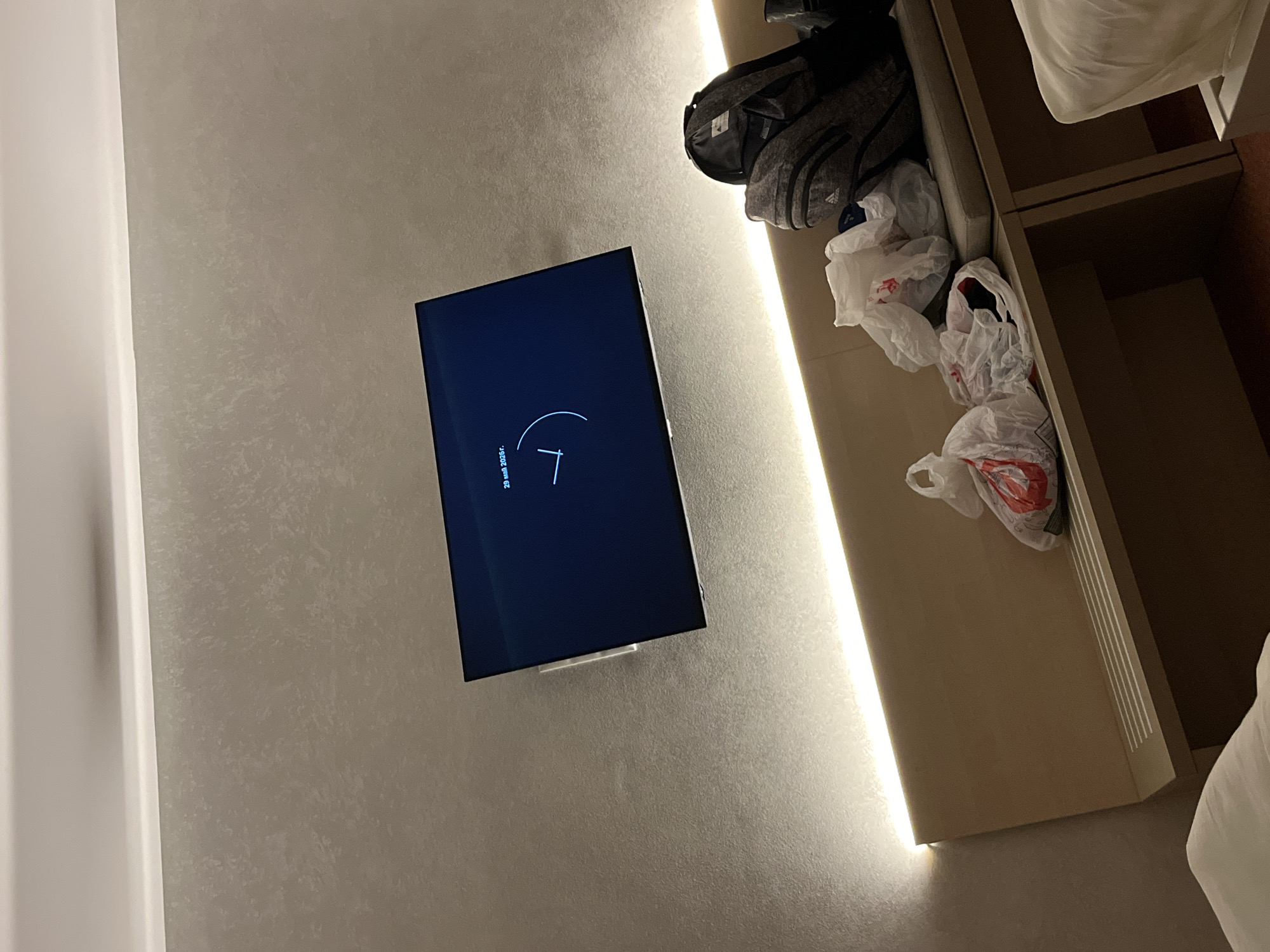 The image depicts a hotel room interior. A flat-screen television is mounted on a textured, light-colored wall. The screen displays what appears to be a clock or a loading animation with the date "29 mai 2026." To the right of the TV, there is a built-in storage unit with open shelves. A gray Adidas backpack sits on the top shelf. Several plastic bags, some bearing a red "R" logo, are clustered on the shelves. Part of a bed is visible, showing white bedding. The lighting in the room is warm, with an accent light strip running vertically next to the storage unit. The photograph provides no verifiable data about the city or location of the image. The description can be given in Belchin, Bulgaria. The photograph provides no verifiable data about the location of the image, including the location in the city of Belchin, Bulgaria.
