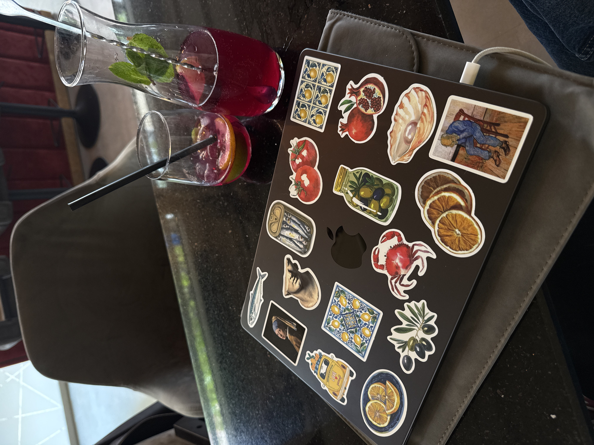 The image presents a laptop adorned with numerous eclectic stickers, placed on a dark tabletop alongside two glasses and a pitcher of a red beverage. The drinks, garnished with mint leaves and fruit slices, suggest sangria or a similar fruit punch. One glass has a black straw, and the pitcher contains a metal stirrer. A white charging cable is plugged into the laptop, which rests on a grey padded sleeve.
The laptop's stickers display a range of illustrations, including pomegranates, a conch shell, tomatoes, a jar of olives, a can of sardines, a fish, a cat's head, dried orange slices, a red crab, an olive branch, and a bowl of sliced citrus. Art-themed stickers feature a scene reminiscent of Van Gogh's "Prisoners Exercising," a depiction of "Girl with a Pearl Earring," and patterned tiles with lemons. A yellow car sticker also shows the word "LOVE."
The setting is an indoor space, likely a cafe or a relaxed work environment, with natural light suggesting it is daytime. Reflections on the tabletop and hints of outdoor greenery in the background add to the ambient light. A dark chair is partially visible on the left, and a glimpse of blue fabric, possibly jeans, suggests a person is seated nearby. The overall scene conveys a casual, leisurely atmosphere, possibly in Plovdiv, Bulgaria, given the provided context.