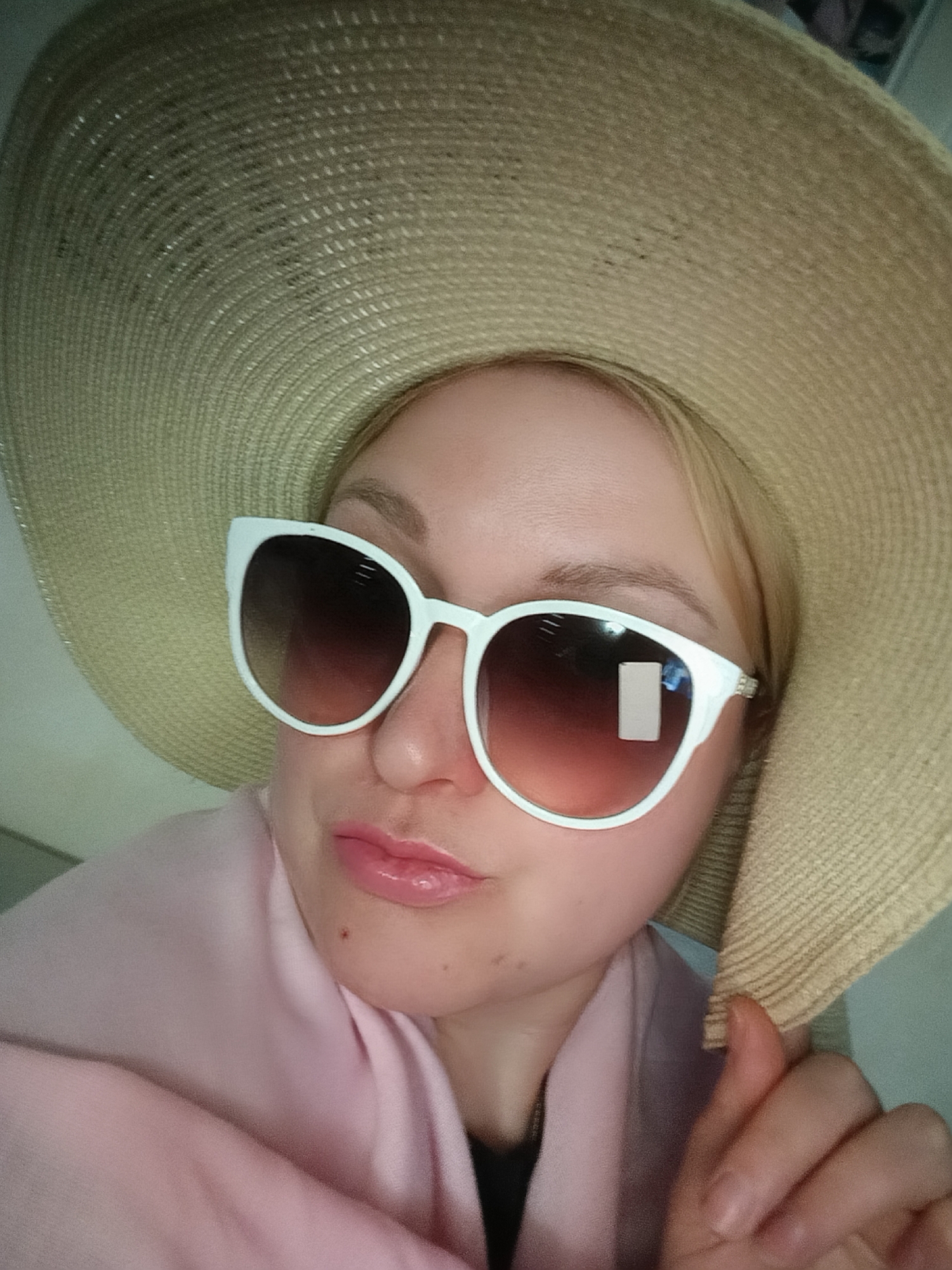 A light-skinned woman with blonde hair wears an oversized straw hat and white-framed sunglasses with gradient lenses. She is also wearing a pink shawl or scarf. A reflective sticker is visible on the right lens of her sunglasses. Her lips are glossy. The image appears to have been taken indoors in Chaikovsky, Russia.