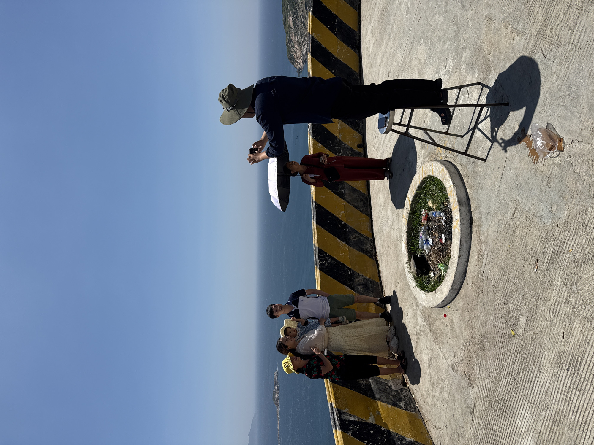 The image depicts an outdoor observation deck in Vinh Hai, Vietnam, on a clear, sunny day overlooking a vast blue ocean with distant islands and landmasses. The foreground features a concrete surface with parallel grooved patterns, indicative of a structured public area. A low, circular concrete planter, approximately 1.5 meters in diameter, is embedded in the pavement, containing dirt, sparse green vegetation, and assorted refuse including plastic bottles and wrappers. A prominent black and yellow striped barrier runs horizontally across the midground, separating the deck from the sea below.
On the left side of the frame, a group of four individuals stands near the barrier, posing. This group consists of an older woman wearing a black floral shirt, black capri pants, and a yellow bucket hat; a younger woman in a light-colored long dress and white clogs, holding a young child in a straw hat; and a man in a white and navy polo shirt with olive shorts. Their gazes are directed towards the distant ocean or slightly to their left.
On the right side, an adult male, facing away from the viewer, stands elevated on a metal stool. He wears a dark long-sleeved shirt, dark pants, and a broad-brimmed khaki hat, holding a black digital camera to his eye, evidently photographing the group on the left. A smartphone rests on the seat of the stool beneath his feet. To his left, an adult female stands, holding a white and black umbrella for shade. She is dressed in a maroon two-piece outfit and carries a small dark cross-body bag. Strong sunlight casts distinct shadows across the concrete. A clear plastic bag containing a brown liquid or substance lies on the ground near the base of the photographer's stool. Scattered small pieces of litter are visible on the concrete surface. Several small boats are discernible on the distant ocean.