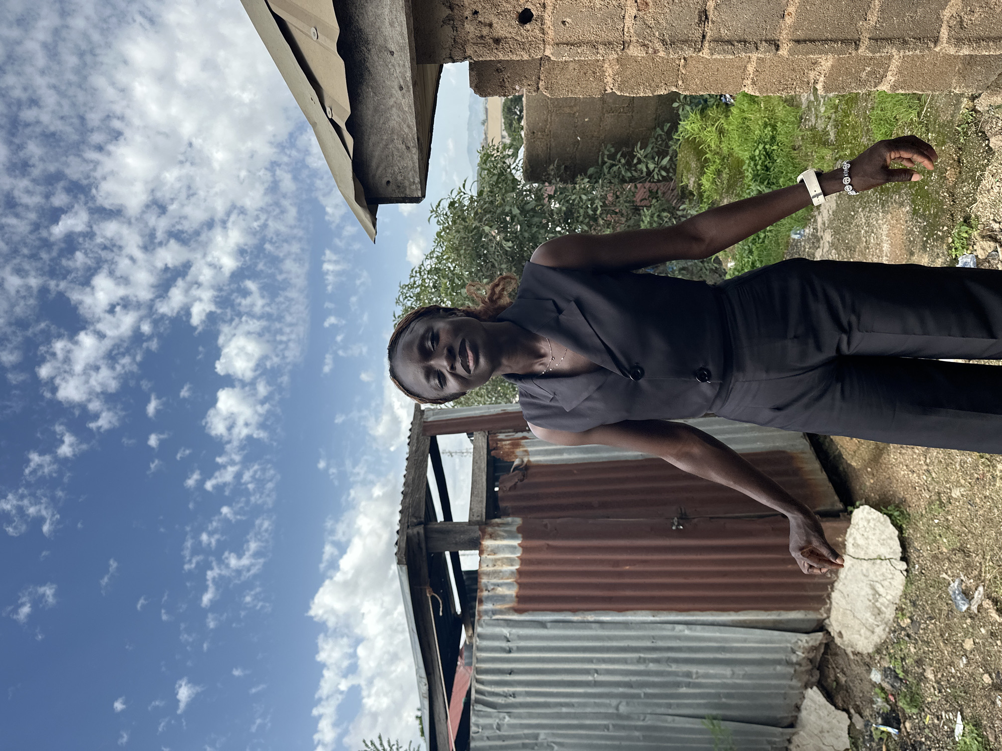 A young woman with dark skin and braided hair stands outdoors, posing for a photo. She is wearing a black vest over a black top and black pants, with a white watch on her left wrist and a delicate necklace. Her head is tilted slightly, and she is looking towards the camera with a neutral expression.
The background suggests an informal setting in Jos, Nigeria. To her left, a cloudy blue sky dominates, with some cumulus clouds scattered across it. A portion of a simple, weathered roof structure is visible in the upper left corner. To her right, a rough brick wall runs vertically, and beyond that, there's lush green vegetation and trees. In the mid-ground, a dilapidated structure made of corrugated metal sheets, some rusted, stands prominently. The ground is a mix of dirt, broken concrete, and scattered debris.
The lighting indicates it is daytime, likely late morning or early afternoon, given the brightness of the sky. The weather appears to be clear with scattered clouds. The overall scene depicts a moment captured in a somewhat rustic and urban fringe environment.