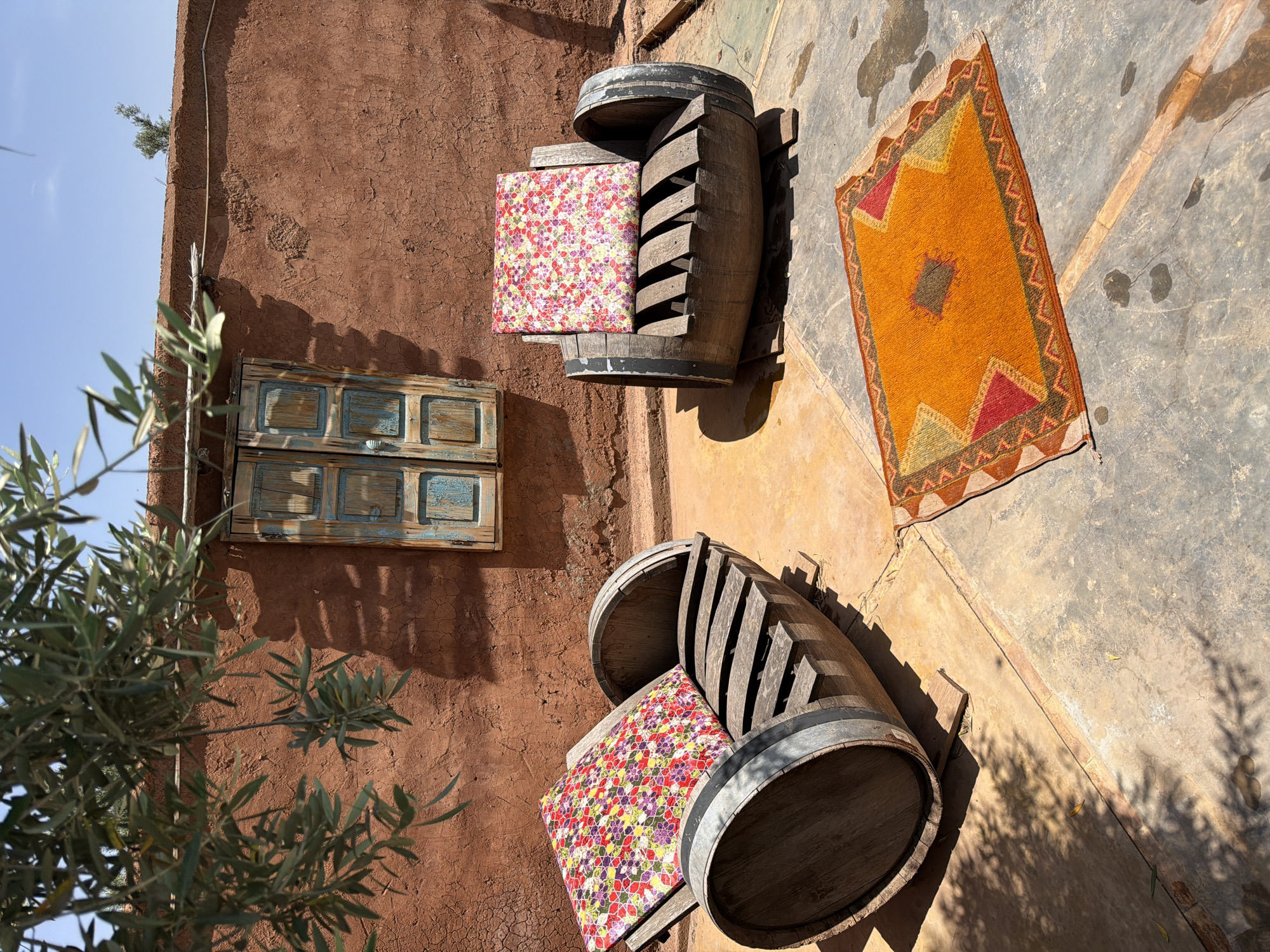 This image captures a vibrant and rustic outdoor seating area, likely in Tahannaout, Morocco, under bright sunshine. The scene is dominated by a textured, reddish-brown earthen wall, possibly adobe or mud brick, which shows signs of weathering and cracking.
In the foreground and midground, two large wooden barrels have been creatively repurposed as seats. Each barrel is cut in half horizontally, with wooden slats forming a backrest, and is adorned with a brightly patterned cushion. One cushion features a colorful floral design with red, pink, yellow, and purple hues, while the other appears similar.
On the greyish concrete or stone ground to the right, a rectangular rug with a warm color palette is spread out. It displays geometric patterns in shades of orange, yellow, red, and dark brown, characteristic of traditional Moroccan textiles. Strong shadows cast by the barrels and overhead foliage indicate bright sunlight, suggesting it is midday or early afternoon.
To the left, olive tree branches with silvery-green leaves partially obscure the view, adding a natural element to the composition. Higher up on the earthen wall, a small, faded wooden window or shutter structure, painted in a distressed blue, adds a touch of aged charm. A thin cable or wire runs along the top edge of the wall, where a small green plant also grows. The sky, clear and blue, is visible in the upper left corner. No people are present, and there is no visible text. The overall atmosphere is inviting, peaceful, and distinctly North African.