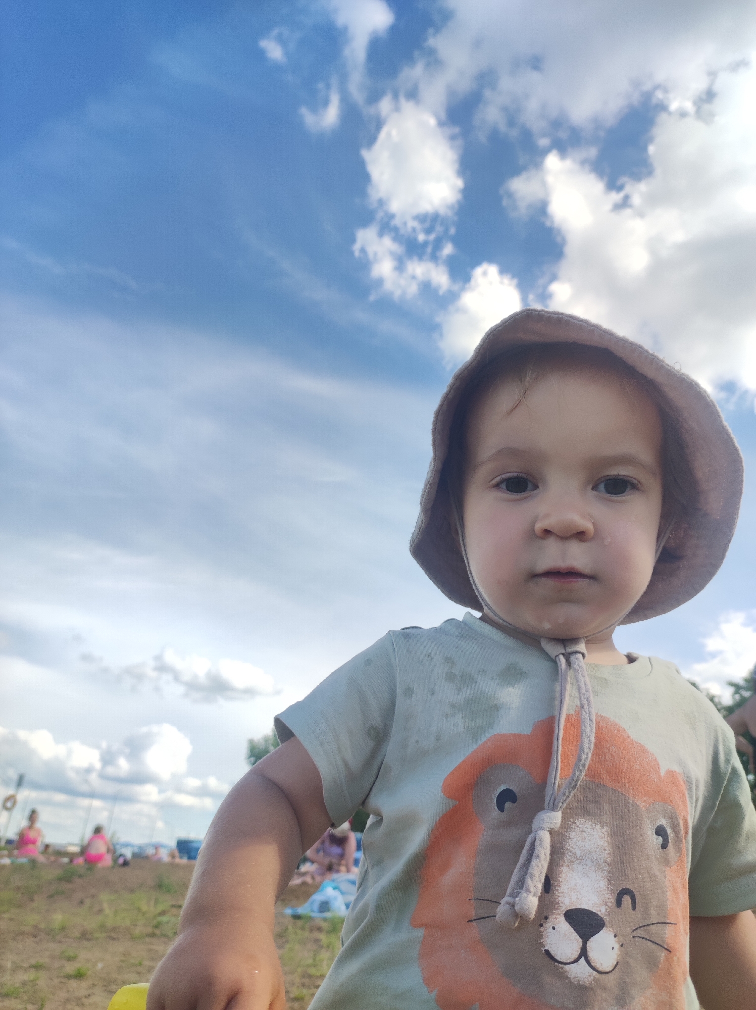 A young child, wearing a light-colored bucket hat and a green t-shirt with a lion graphic, is positioned in the foreground looking directly at the camera. The child's skin appears slightly damp, and there are visible water droplets on their face. Behind the child, the background shows a beach scene with scattered people in the distance, some wearing swimsuits and others sitting on the sand. The sky above is a bright blue with scattered white clouds. The overall setting suggests an outdoor recreational area, likely a beach or park, during daylight hours. The image does not contain any specific elements that would indicate it is located in Chaikovsky, Russia.