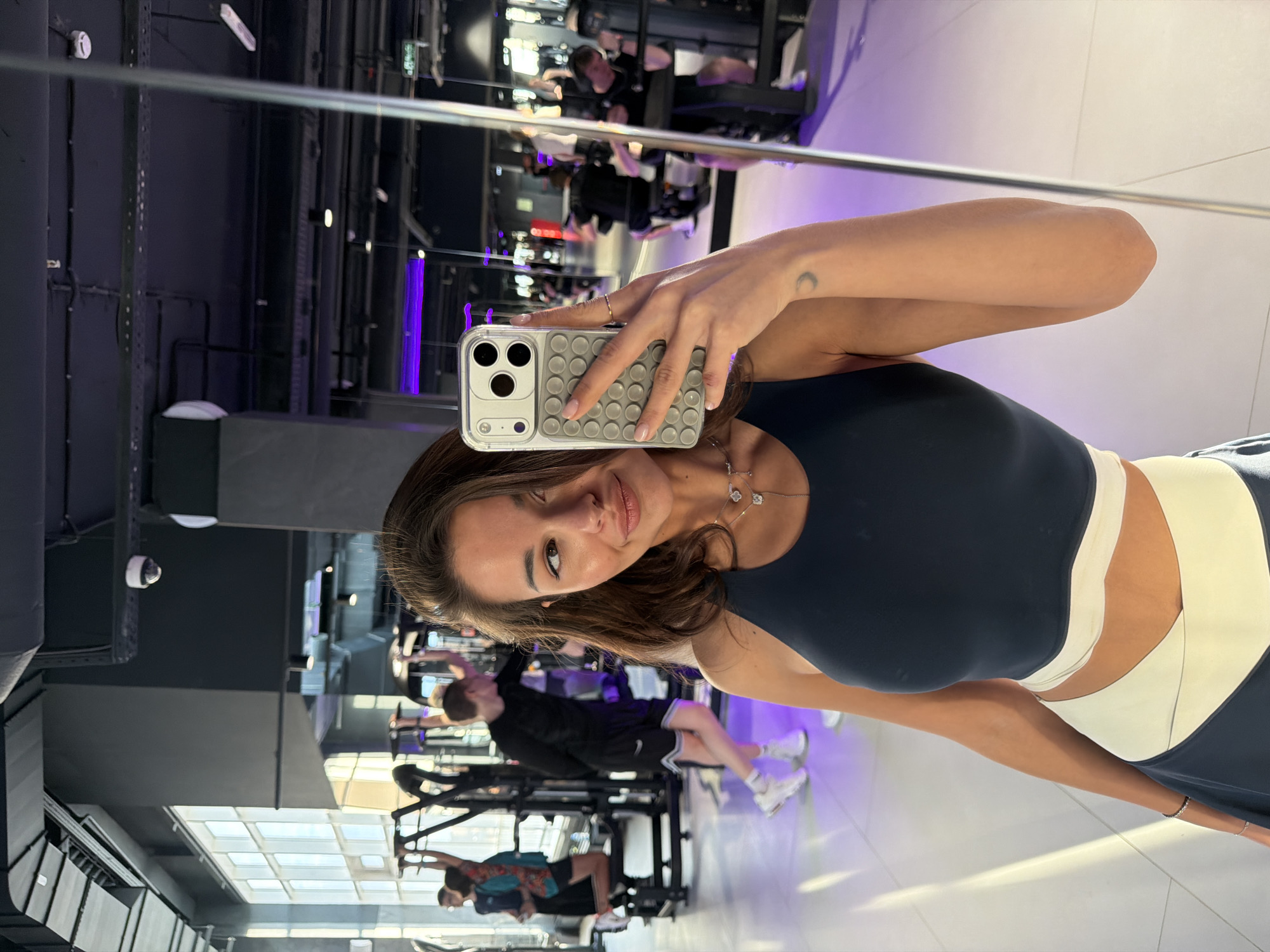 The image captures a woman taking a selfie in a gym setting. She is holding a smartphone with a light grey patterned case. The gym environment is visible in the background, with workout equipment and other individuals exercising. The woman is wearing athletic clothing, specifically a dark top with light-colored accents. The location is identifiable as the World Class Krasnoyarsk gym in Krasnoyarsk, Russia, based on the gym's interior design and layout.