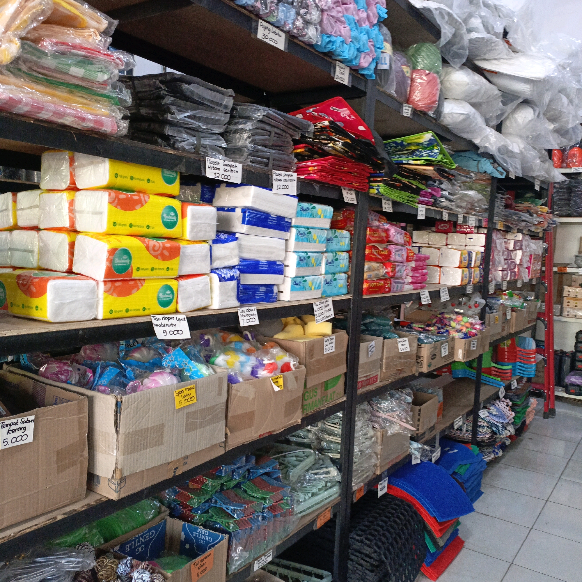 The image depicts a retail store in Salatiga, Indonesia, specializing in household goods and plastic products. The scene is dominated by shelves stacked with various items, primarily packaged goods such as tissues, plastic bags, and cleaning supplies. Several shelves are filled with neatly arranged stacks of boxed and individually wrapped products. Prominently displayed are numerous packages of facial tissues and kitchen towels, some with bright yellow and orange packaging. Other items include rolls of green and pink rope, a variety of plastic containers, and stacks of colorful doormats. The store appears to be well-stocked, with a dense arrangement of merchandise on metal shelving units. Cardboard boxes are also visible, some used for storage and others containing goods. Prices are indicated on some of the items with handwritten tags. The overall environment suggests a busy, functional retail space.