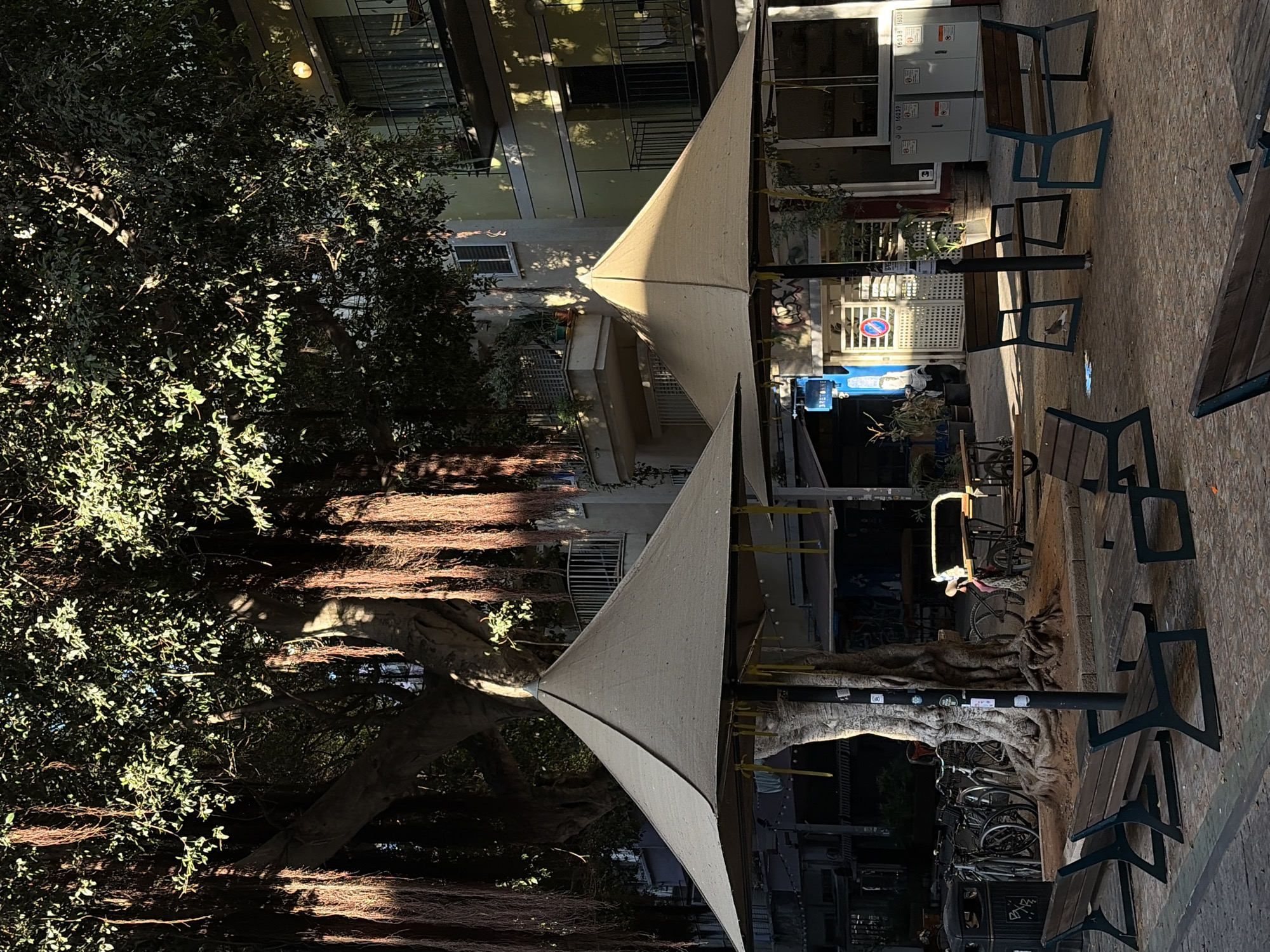 This outdoor shot captures a shaded corner of Tel Aviv, Israel. Tall buildings and a leafy tree dominate the background. The outdoor seating features wooden benches and tables set up around the tree. Fabric shades overhead provide sun protection. Bicycles parked in the shadows are noticeable, adding a local vibe to the scene.