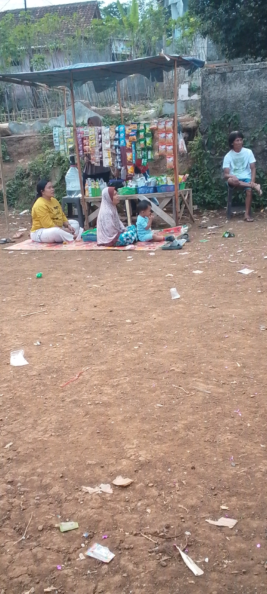 In Cilaku, Indonesia, an outdoor scene unfolds near a roadside stall laden with snacks and drinks. Three individuals are seated on the ground in front of the stall. To the left, a woman in a yellow shirt and white pants sits cross-legged on a mat. Beside her, a woman in a headscarf and a patterned blue and white skirt sits with her legs tucked to the side. A young child, dressed in a blue outfit, sits between them, facing away from the camera. A man in a white t-shirt and blue shorts is seated on a stool to the right, facing the stall. The ground is composed of dirt, with various pieces of litter scattered around, including plastic cups, wrappers, and what appears to be a red string. Behind the stall, a simple building with a tiled roof and trees are visible, suggesting a semi-rural or suburban environment.