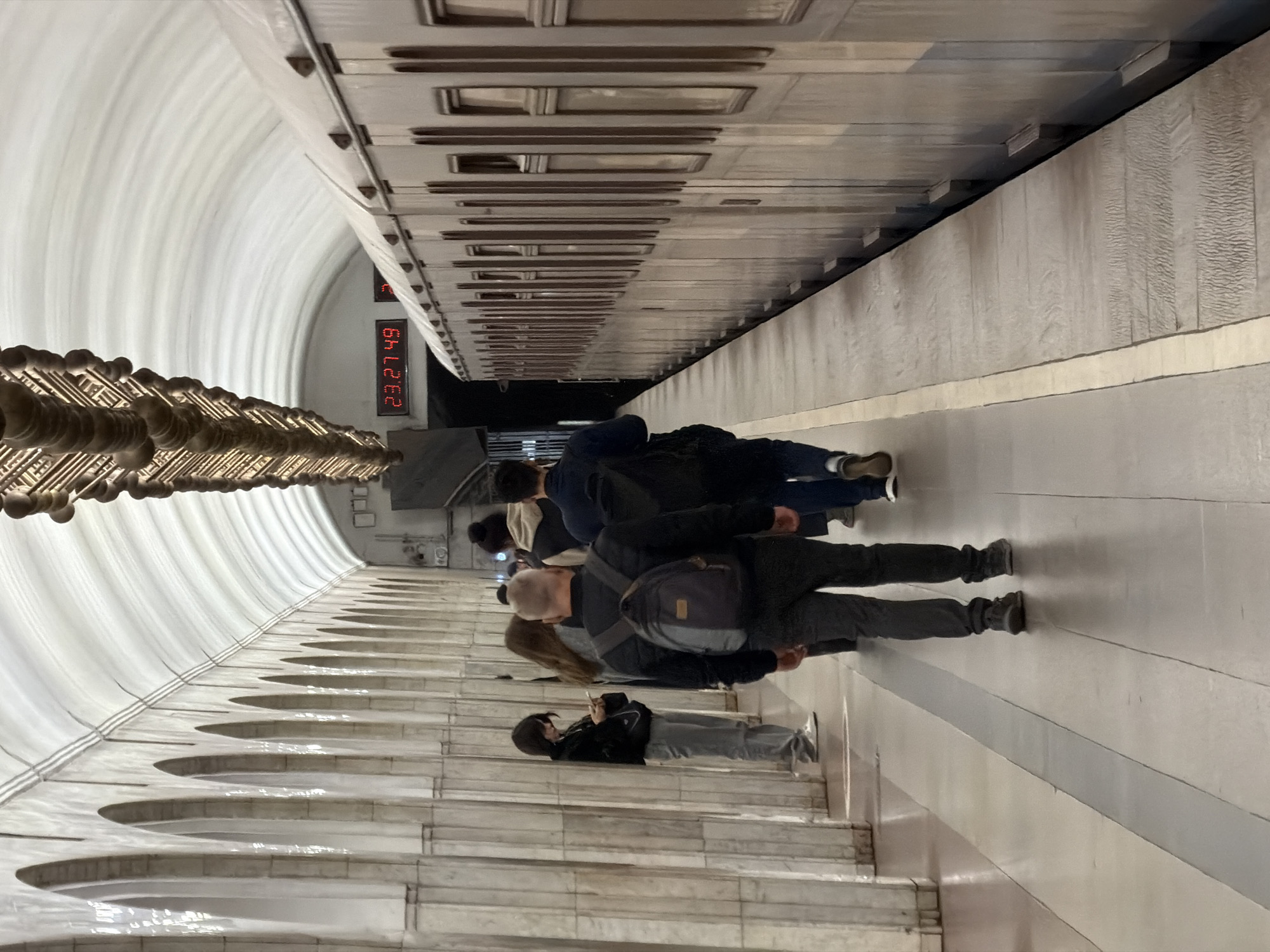 The image captures a scene in the Moscow Metro, Russia. A subway train is stationed at a platform. Several people stand on the platform waiting for the train. A large digital clock displays the time, "23:27:49". The station's architecture includes arched walls and ornate lighting fixtures. The overall environment is brightly lit.