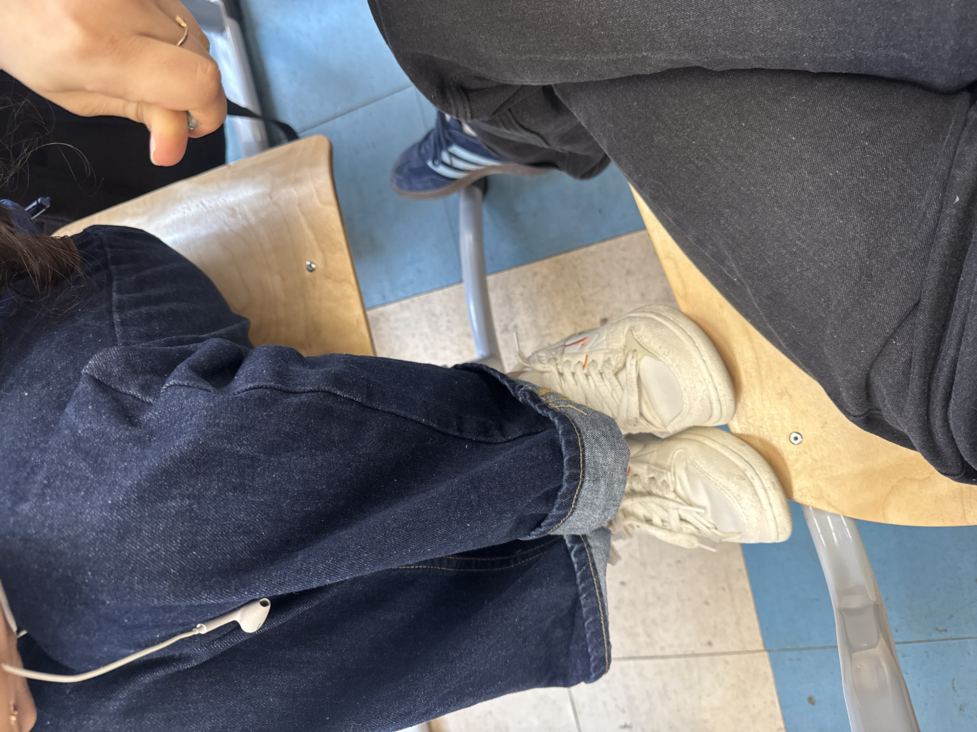 Two people are sitting in classroom chairs. One person is wearing dark blue jeans, rolled up at the cuff, and white sneakers with orange detailing. The second person is wearing dark grey jeans and blue sneakers with white stripes. One person has a ring on their finger and an earbud cable is visible. The chairs have light wood seats and blue and white metal frames. The location is indoors. There is no definitive information that suggests this image was taken in the city of Saint Petersburg, Russia.