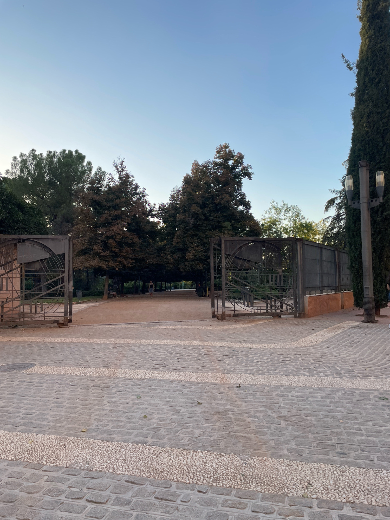 The image depicts an entrance to a park or garden in Granada, Spain. In the foreground, there is a cobblestone pathway with decorative strips of small, smooth white pebbles. The pathway leads to two large, ornate metal gates, one on the left and one on the right, which are open. Beyond the gates, a dirt path winds through a landscape of trees. A single person, appearing to be a woman wearing dark clothing, is walking on the path in the distance, towards the left of the frame. A lone figure is also visible sitting on a bench under a tree in the middle ground. A tall lamppost with two lights stands to the right of the right gate, beside a tall, dark green evergreen tree. The sky above is clear and a pale blue, suggesting it is either early morning or late evening.