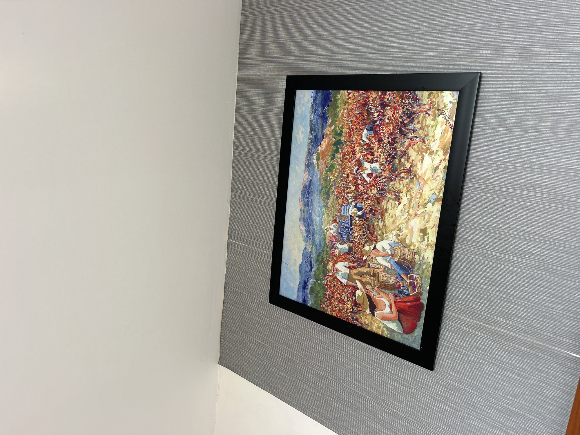 A framed painting hangs on a gray textured wall adjacent to a white wall in an indoor setting. The painting depicts a scene of people working in a field, possibly a vineyard or other agricultural setting. The painting is predominantly red, orange, and blue, suggesting an impressionistic style. The location is Bac Nha Trang Ward, Vietnam.
